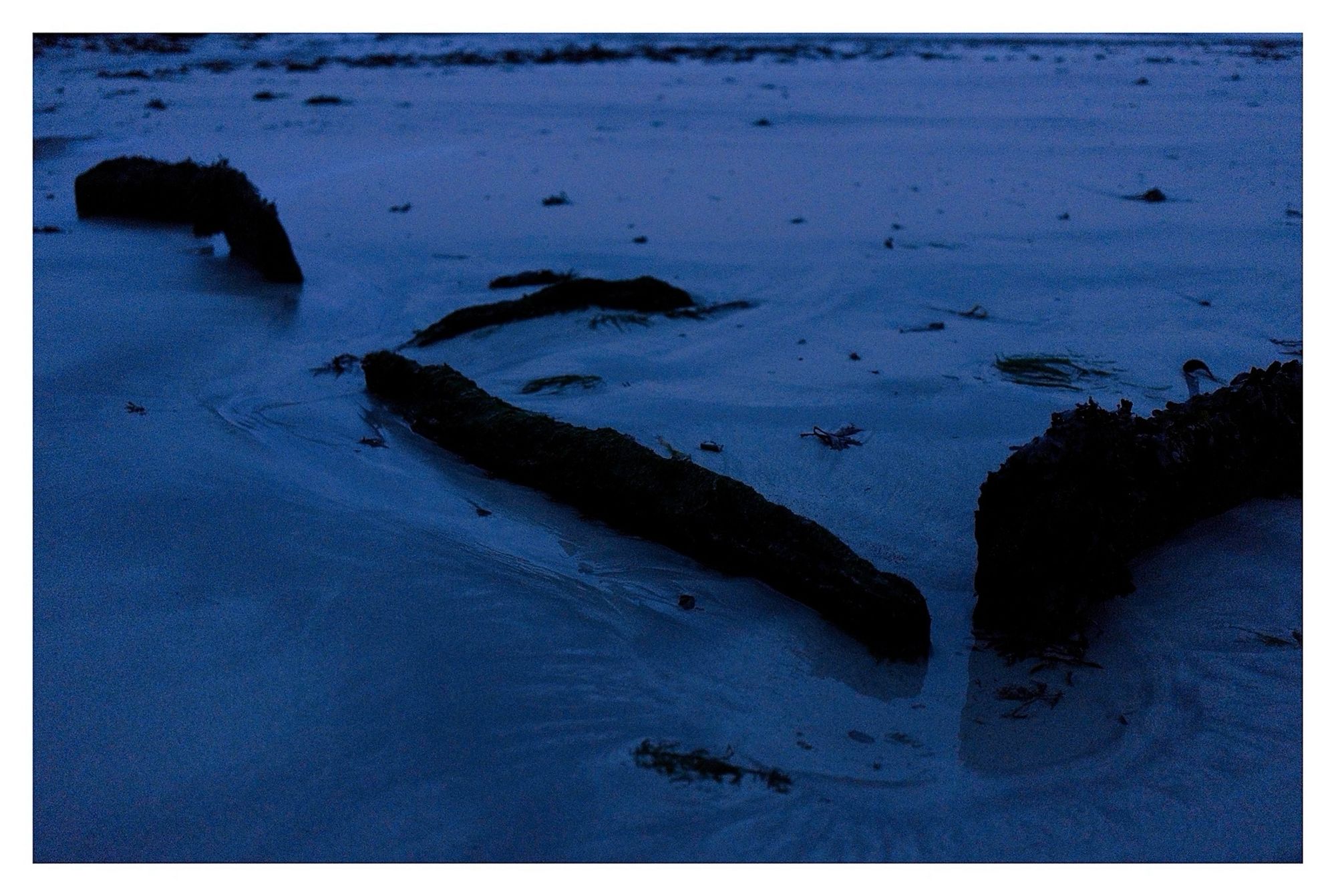 Description: On mid-foreshore at low water on S side of Ballyrisode
beach/strand (inner beach) to immediate SE of carpark; site would cover at mid to high water. Eroded and well buried, so difficult to assess nature and extent. 2 upright slabs at slight angles to each other facing seaward and may have been a third stone but is now collapsed; 1m to south 3 long slabs enclosing a rectangular box-like stone setting; enclosed on 3 sides and open on side facing seaward. May be an earlier site reused for later burials. Potential fulacht fiadh stone trough or a cist burial.