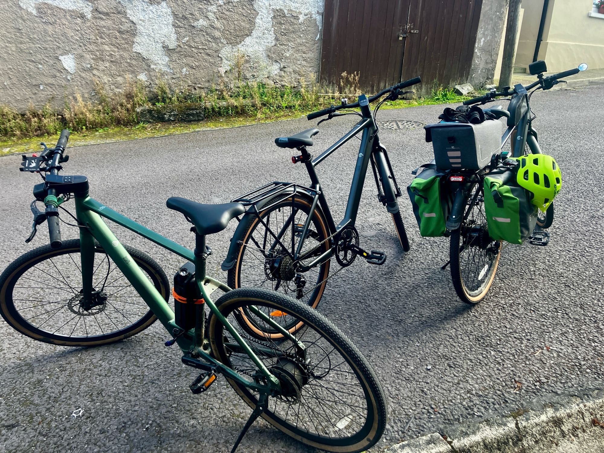 Three eBikes on a hill. 

These are the same make and model bike. 

One a large frame in green, the first in the village has a slightly different interface and handlebar controller. The other two, newer, are medium frames in dark grey.

Mine has the panniers, trunk box because I can’t help but carry stuff. Snacks, stove, togs, towels and rain gear. I like to stay out as much as possible without the extras on my back if I can help it.