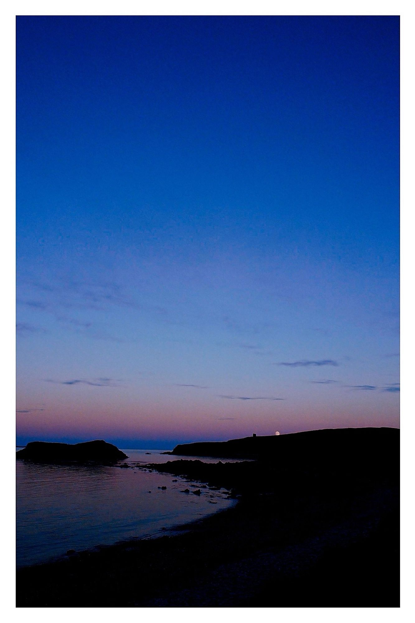 A early moon rises over Horse Island and in to the blues and pinks of a sunset sky. The same colours reflected in the sea along a silhouetted coast line.