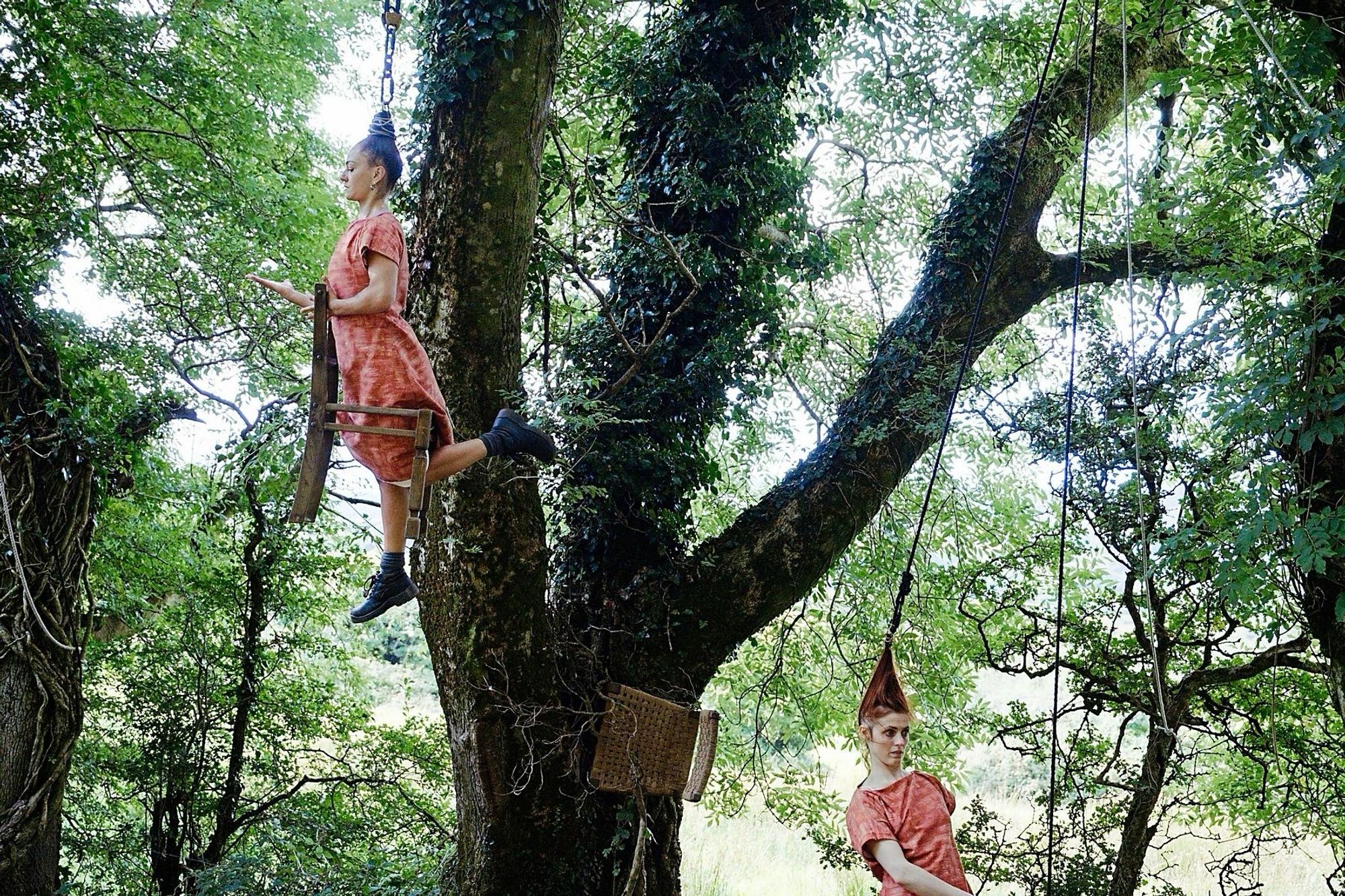 Two performer, Lou (upper left) and Imogen (lower right), both wearing identical red dresses. 

Lou is suspended on a rope by her hair with one knee on a chair she’s holding up with her suspended about 10 feet off the ground. 

Imogen is on the ground her hair pulled up towards the branches also by rope.