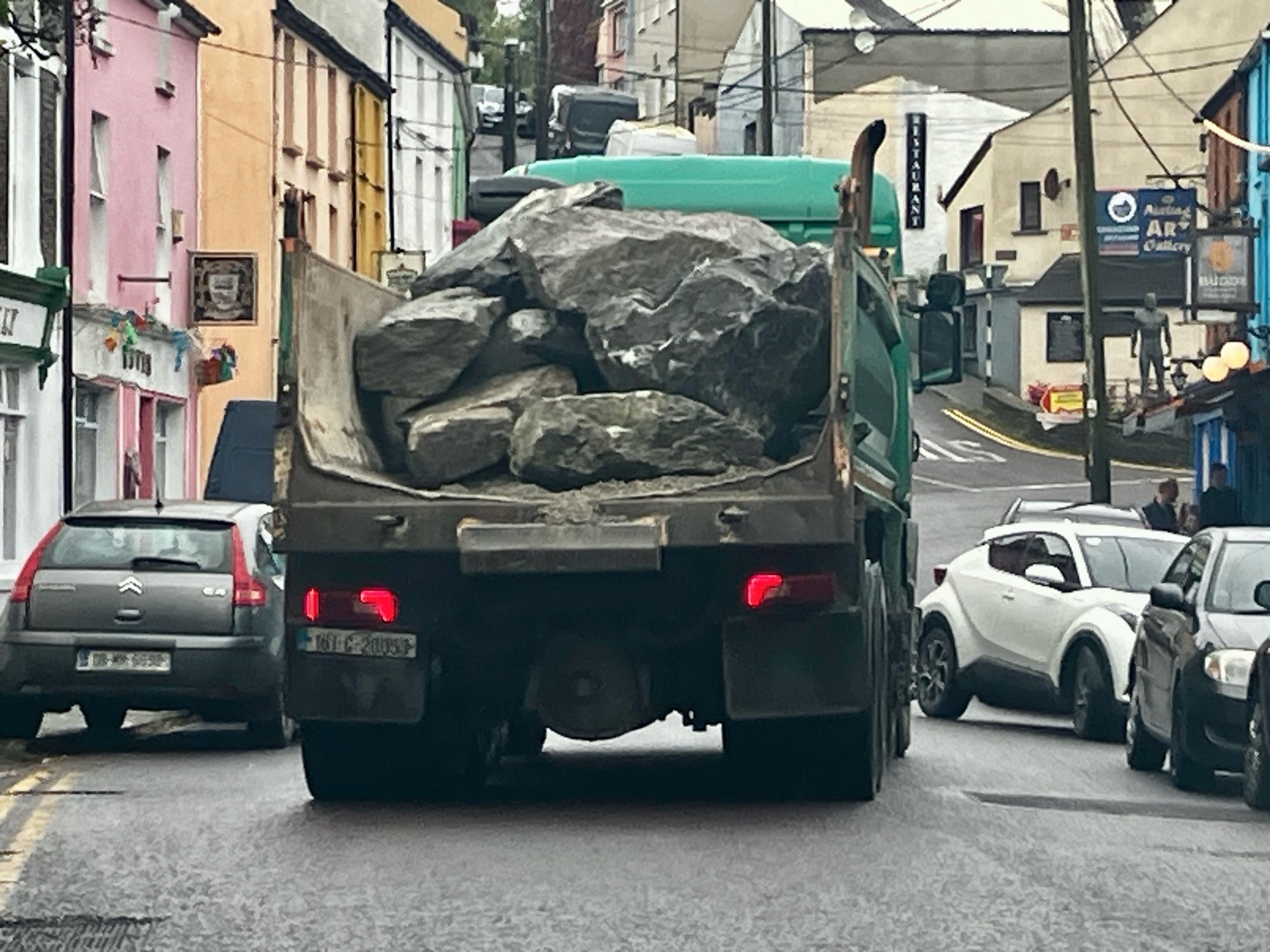 A big truck carrying boulders in an open ended bed heading through a village towards a choice of two steep hills.