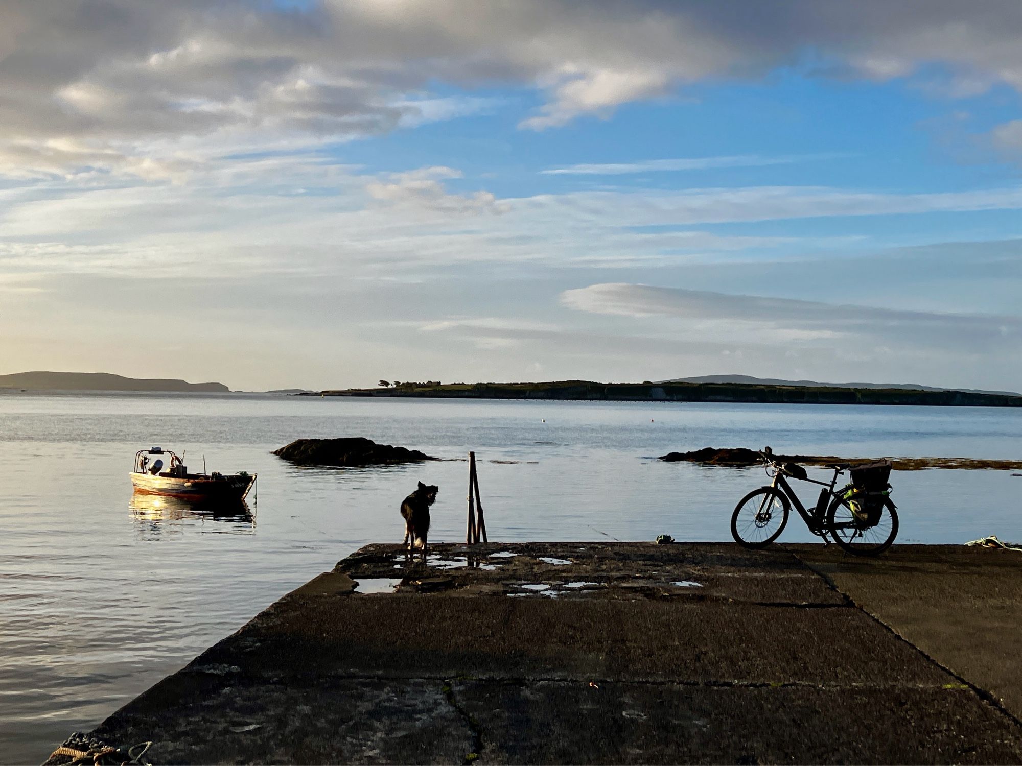 Small pier at sunrise with a dog and bike silhouetted against the sea. A small boat moored nearby and islands on the horizon