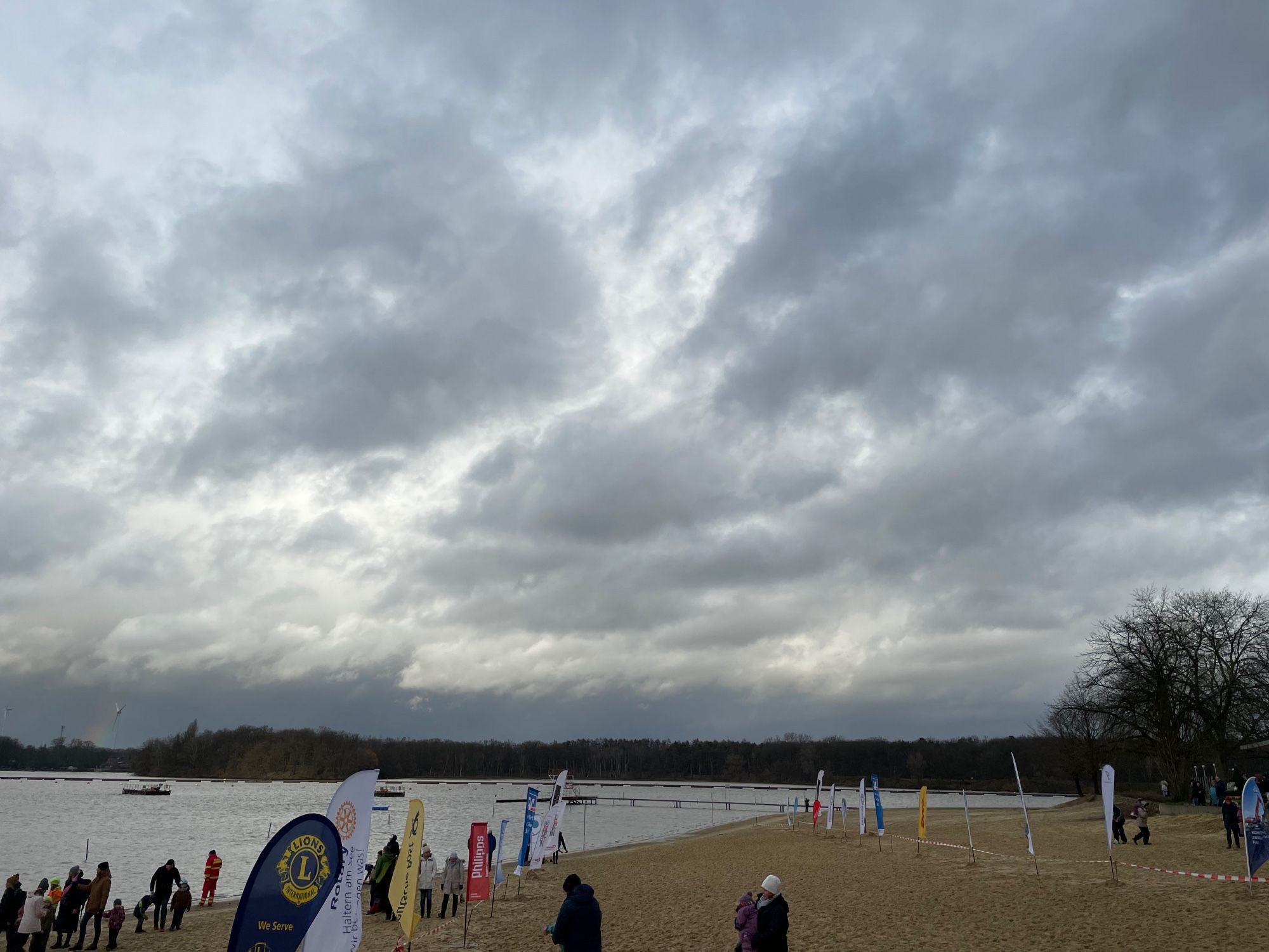 Strandbad mit ein paar Menschen am Ufer, der Himmel ist bewölkt, am Strand stehen Flaggen