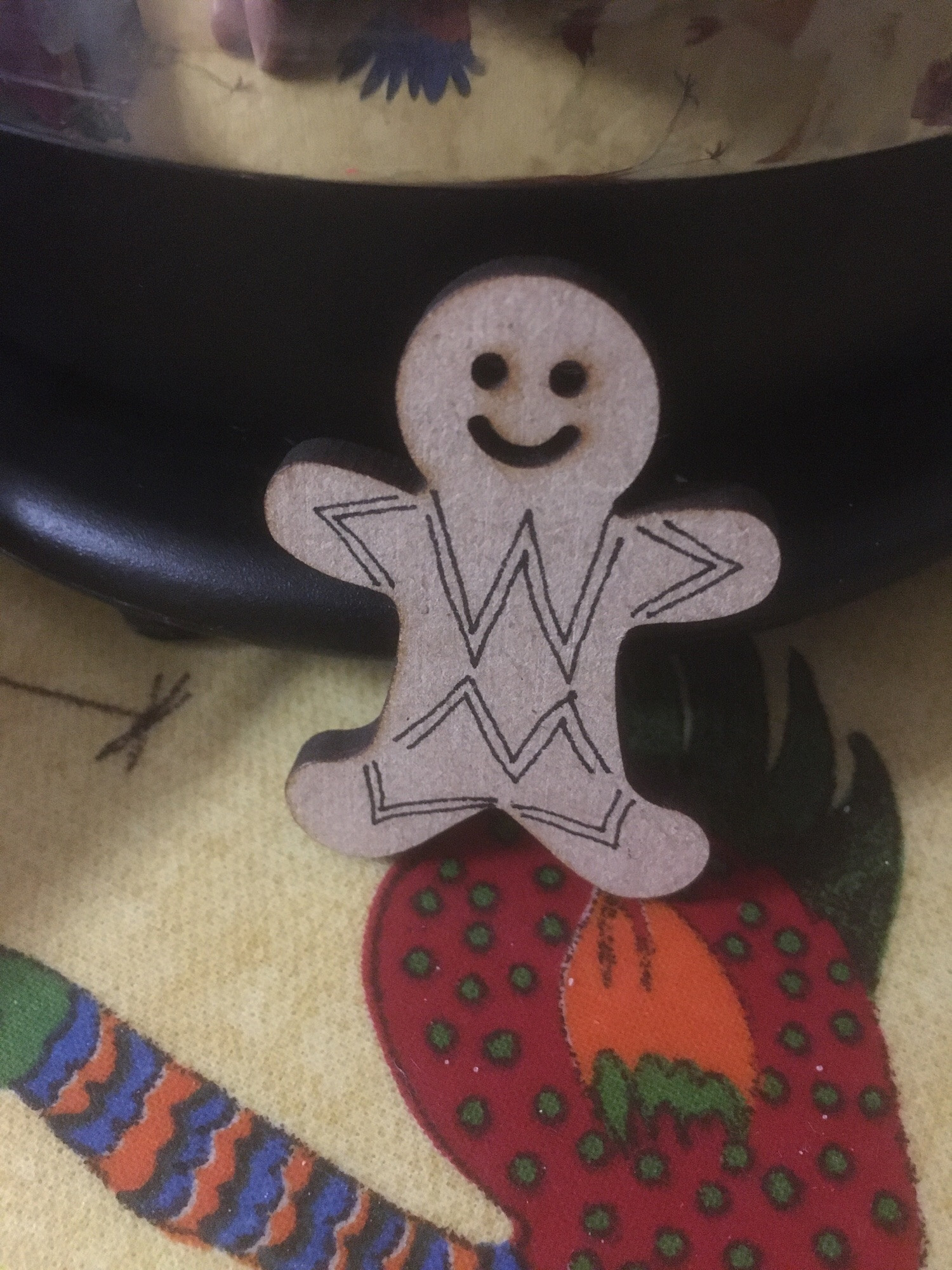 Photo of a small wooden gingerbread man, about five centimetre tall, with diamond patterns drawn on in black ink. He stands on a colourful surface, leaning against the bottom of a shiny kettle.