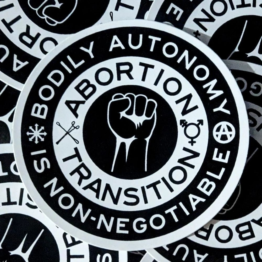 Circular stickers with a white line drawing of a raised fist on black in the middle. In a white border surrounding that are the words “Abortion” and “Transition” in black, accompanied by the trans/gender inclusive symbol and an image of forceps and curettes, the tools used in a dilation and evacuation abortion procedure, crossed in an X shape. In a black border around that, white text reads “Bodily Autonomy is Non-Negotiable” accompanied by the chaos star (eight arrows pointing outwards in a radial pattern from one central point) and an anarchy symbol.