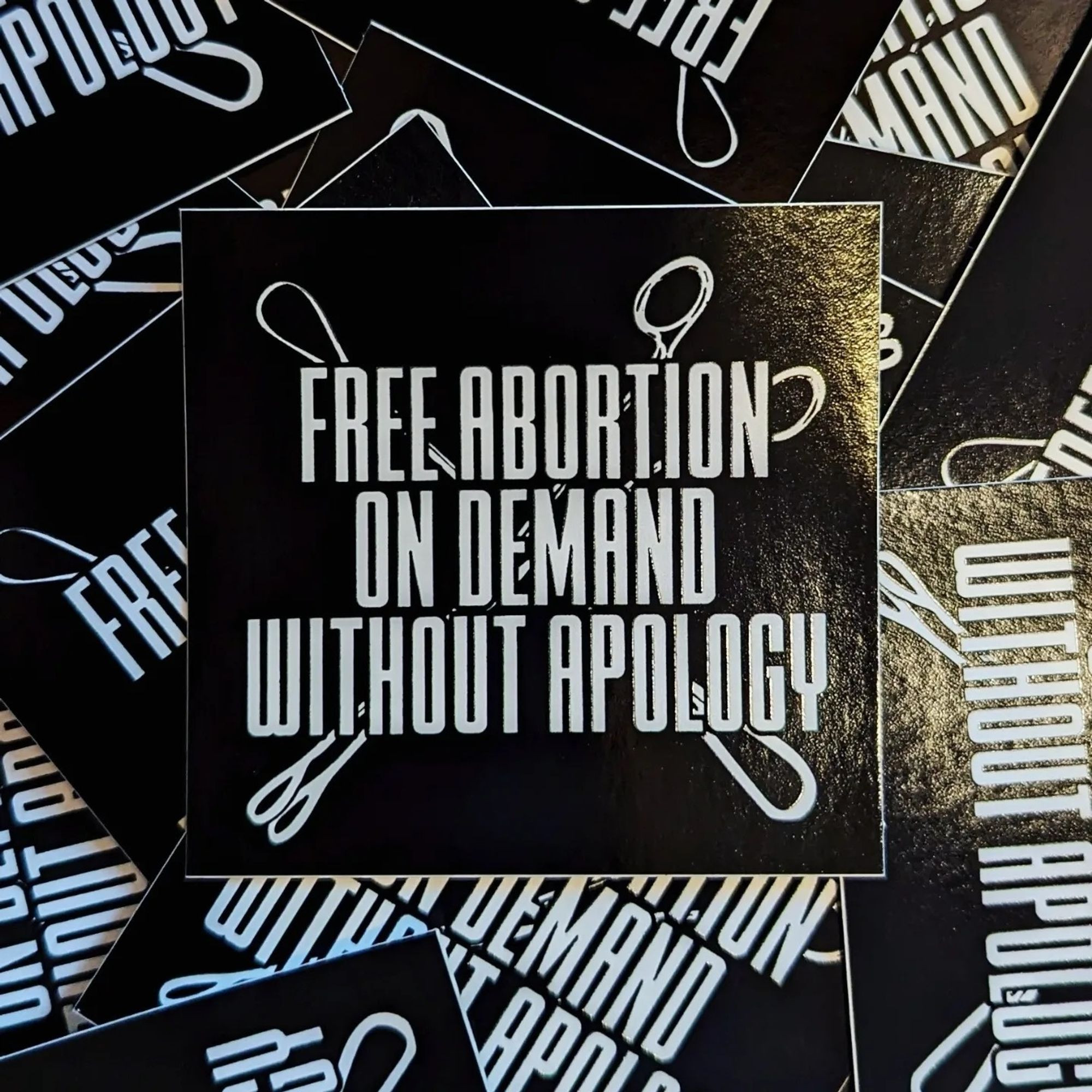 Black stickers with images of white forceps and curettes, the tools used in a dilation and evacuation abortion procedure, crossed in an X shape in the background. In bold white font on top of the image, text reads “Free abortion on demand without apology.”