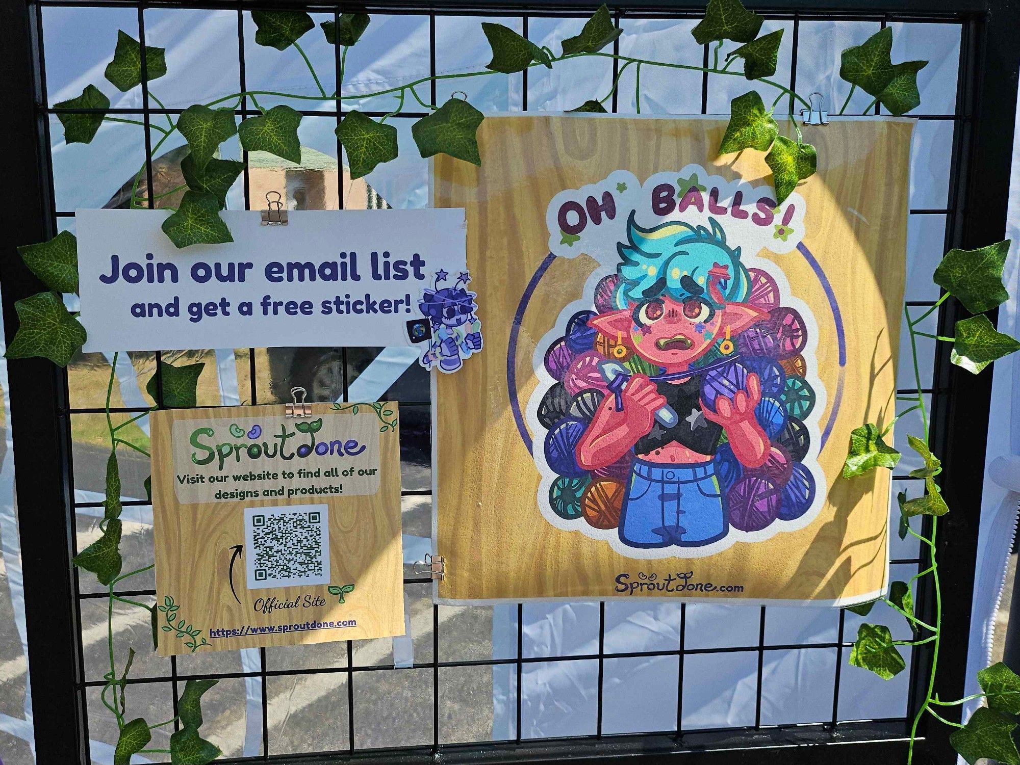 A standing display features three signs.  The largest one is of Sproutdone's Oh Balls sticker with an elf with bright red skin and cyan hair looking exasperated in front of a pile of yarn balls.  The other signs show a QR code for Sproutdone.com and an ad that says, "Join our email list and get a free sticker!"  Next to it is the alien sticker from the previous image.