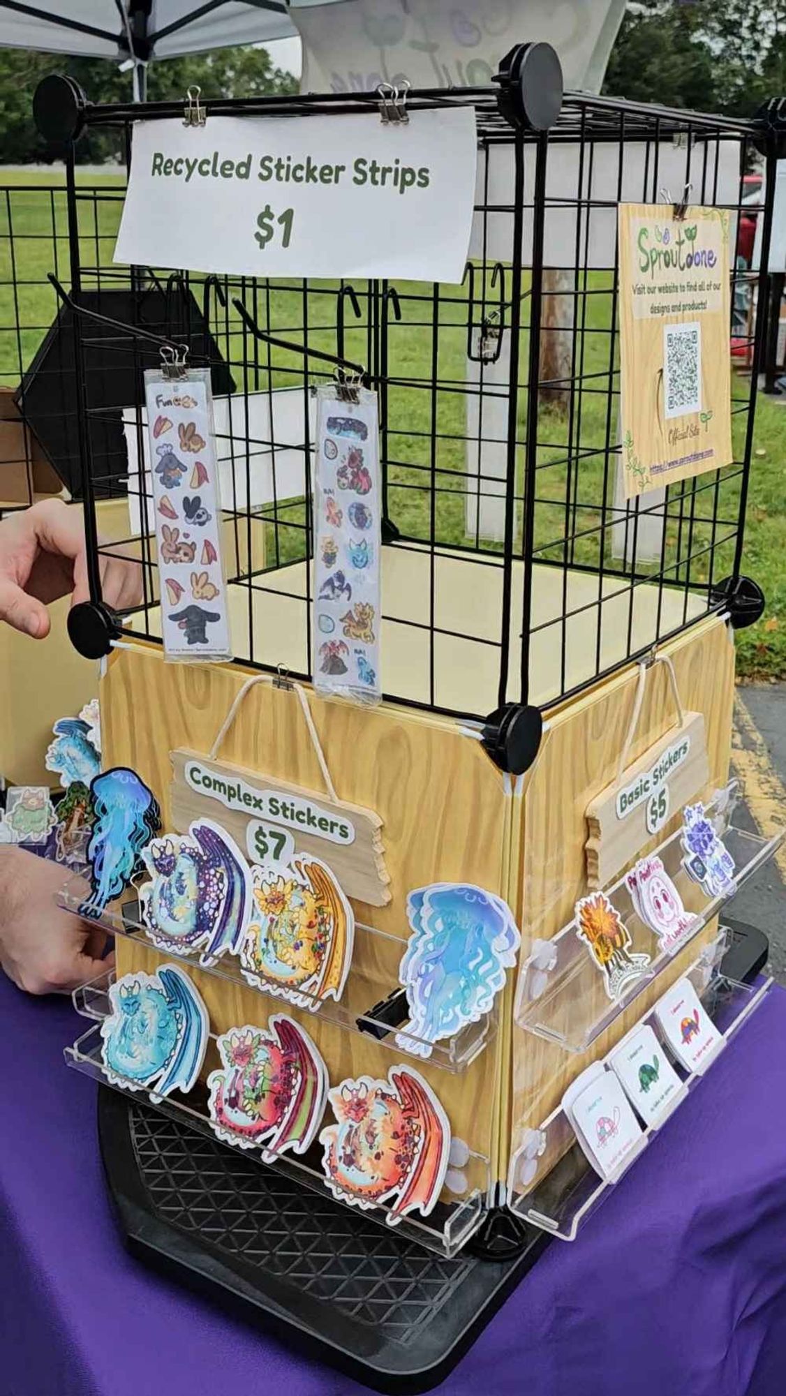 A custom-made spinning display with a cube of grid panels atop a cube of solid panels with a fake wood design.  $1 two designs of recycled stickers strips are featured, including rabbits and dragons.  Below on clear shelves are stickers featuring a humanoid jellyfish, five different colored dragons with babies in eggs, a bee with fall leaves for wings, an alien with glowsticks, and three turtles.