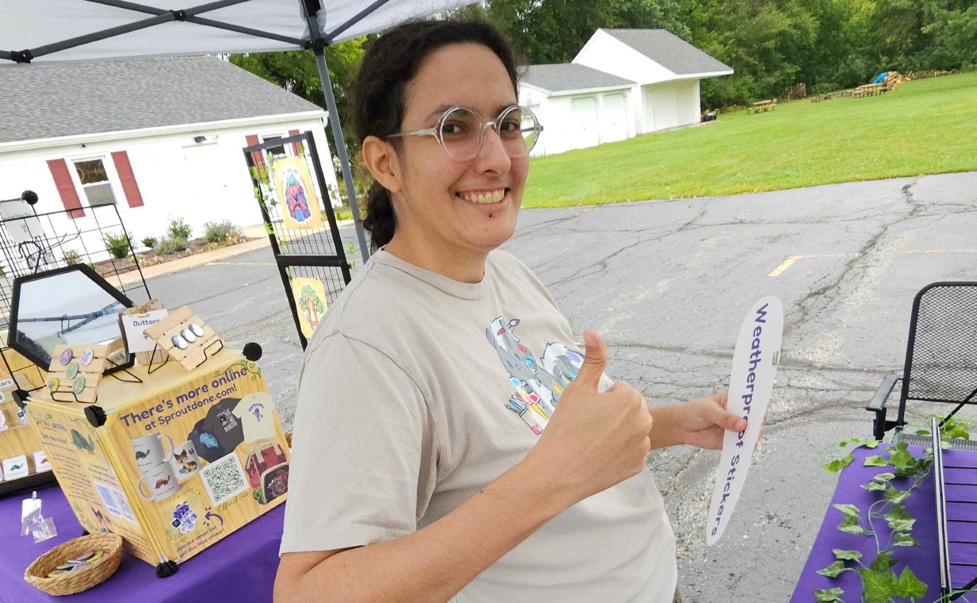 Beans is doing a thumbs up while holding a thin sign that says "Weatherproof Stickers" during setup.  Behind them is their button display and a mirror.  The panel on the cube display says, "There's more online at Sproutdone.com!"  It shows mugs, shirts, and backpacks that are only available for purchase online.  A standing display featuring their artwork is further in the background.