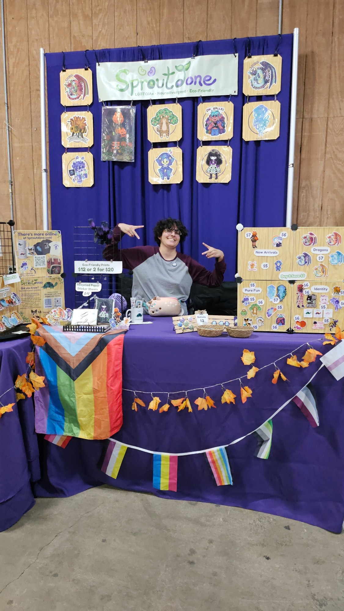 Sproutdone's indoor booth has a purple table with pride flags, brightly colored anime-inspired designs, stickers, prints, and button pins.