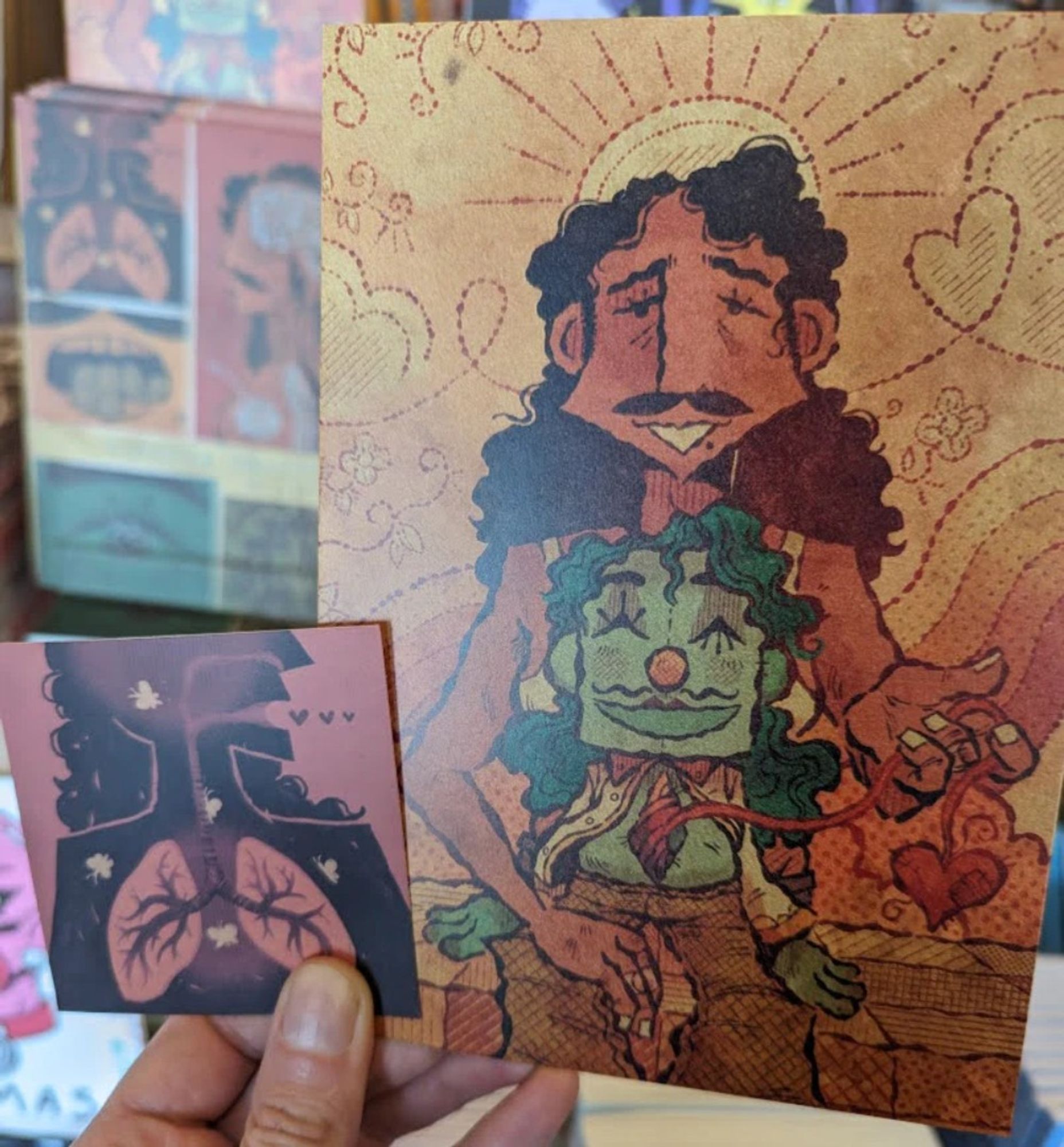 A hand holding up a Viscera Objectica postcard and sticker, with the book blurred in the background.