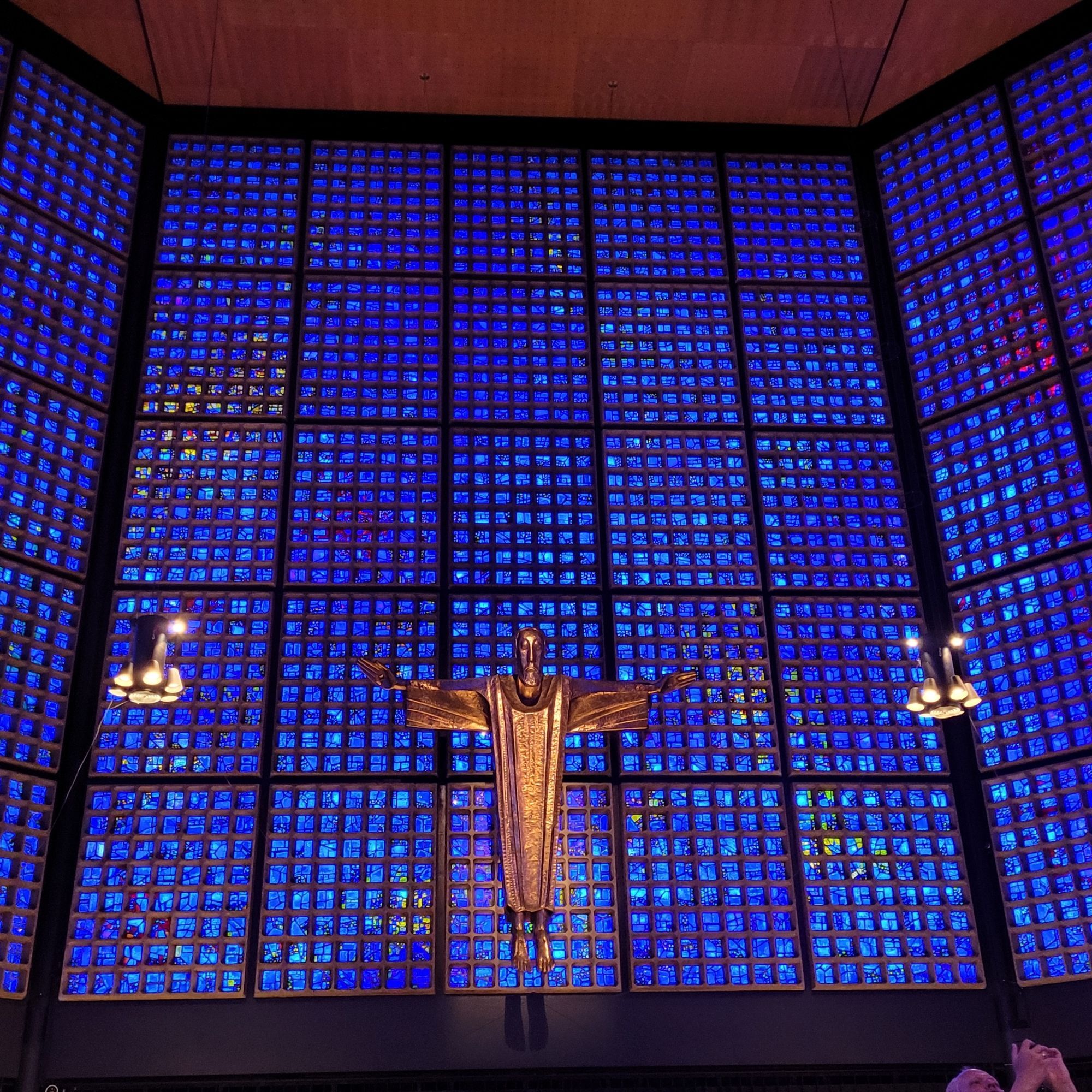 Kaiser Wilhelm Memorial Church
Kaiser-Wilhelm-Gedächtniskirche

An interior view of the second, working church.
There is a wall of blue glass bricks that allow natural light in. Mounted in front of the blue glass wall, there is a large golden figure of Christ in a modern style. This was an incredibly serene space.