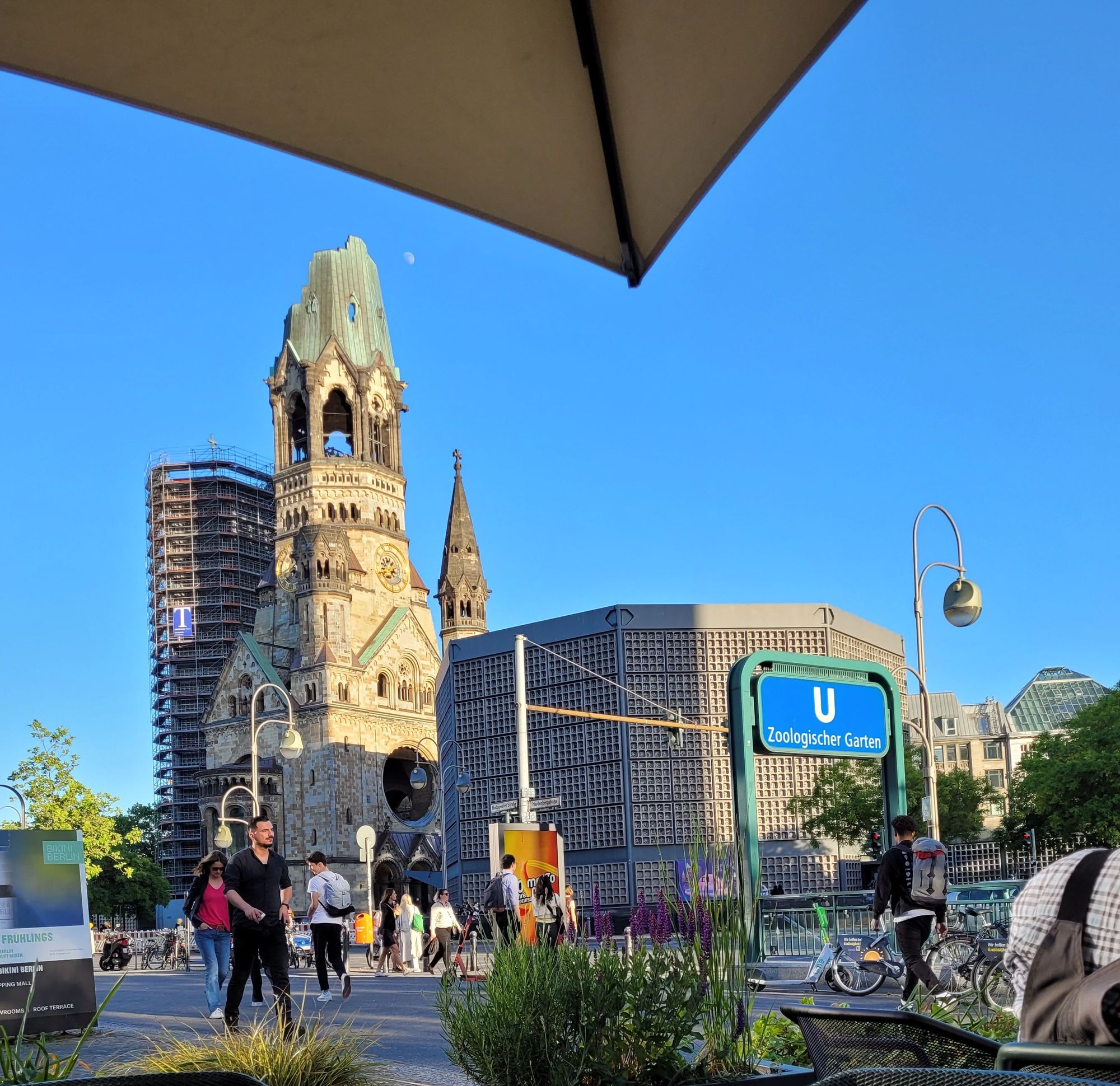 Another picture of Kaiser Wilhelm Memorial Church/Kaiser-Wilhelm-Gedächtniskirche from about a block away. There is a modern bell tower in the same style as the blue church, the damaged original church in the middle, & the blue church to the right. Also visible, pedestrians & a subway sign.