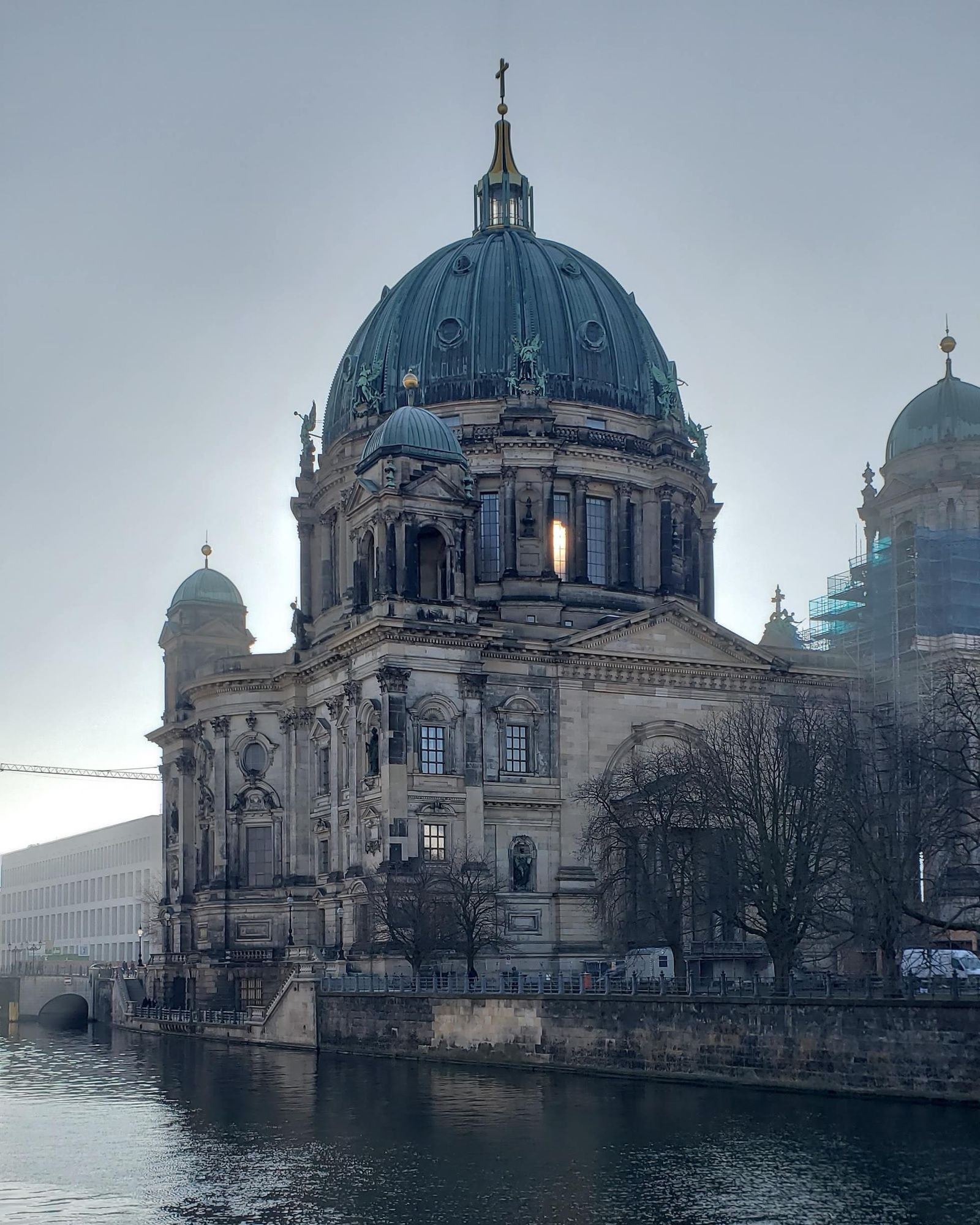 The rear of the Berliner Dom, with the River Spree in the foreground. A light shines through a window facing the camera. 
The belltowers were undergoing renovations in 2019 and are covered in scaffolding.