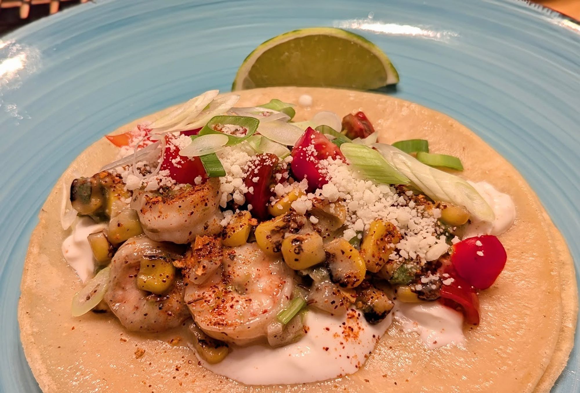 A light blue plate with yellow corn tortillas.  On the tortillas is sour cream, topped with creamy fried corn, shrimp, tomatoes, scallions, and cotija cheese.  There is a lime wedge in the background.