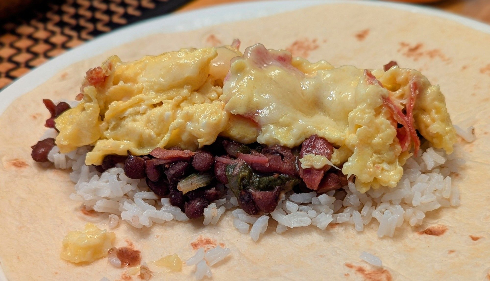 A white plate with a flour tortilla. It is topped with white rice, braised beans and smoked turkey, and cheesy scrambled eggs.