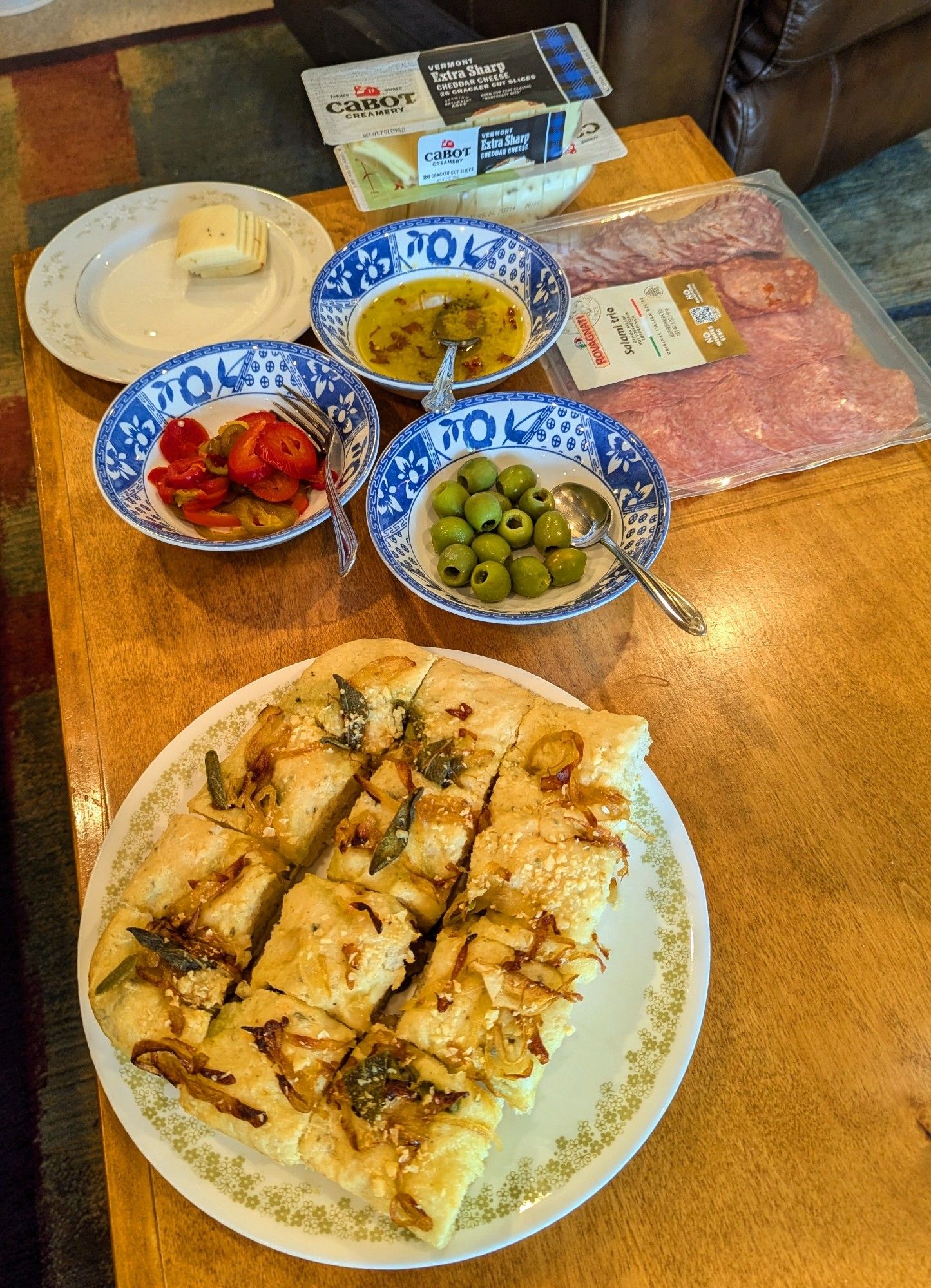 A pine coffee table, stained light brown. A Corelle plate (white with a green floral border) with focaccia topped with onions, sage, and Parmesan. In the background are blue and white bowls with hot, pickled peppers, olives, olive oil and garlic, and assorted salamis and cheese. There is a white plate at 11 o'clock with a small stack of pepper jack cheese.