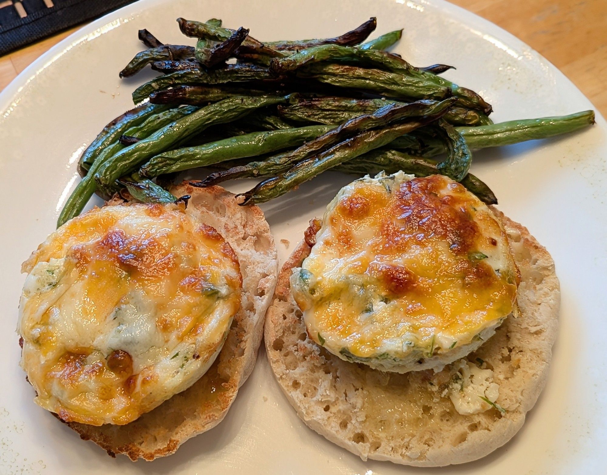 A white plate. 2 toasted English muffins topped with cheese and herb baked eggs. Above those on the plate are a pile of roasted green beans.