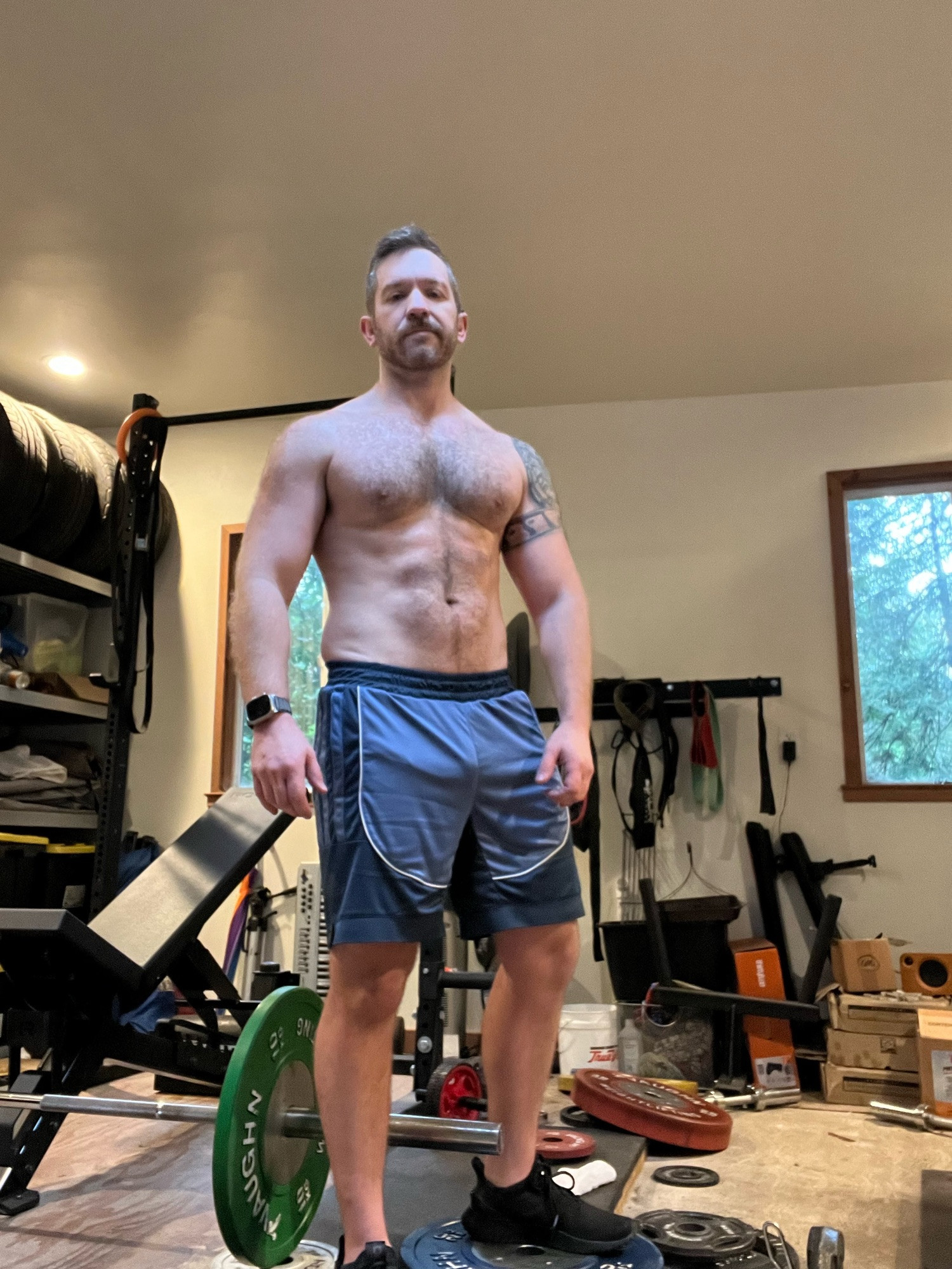 Boss shirtless amid gym equipment in a garage, with redwood trees visible through the window