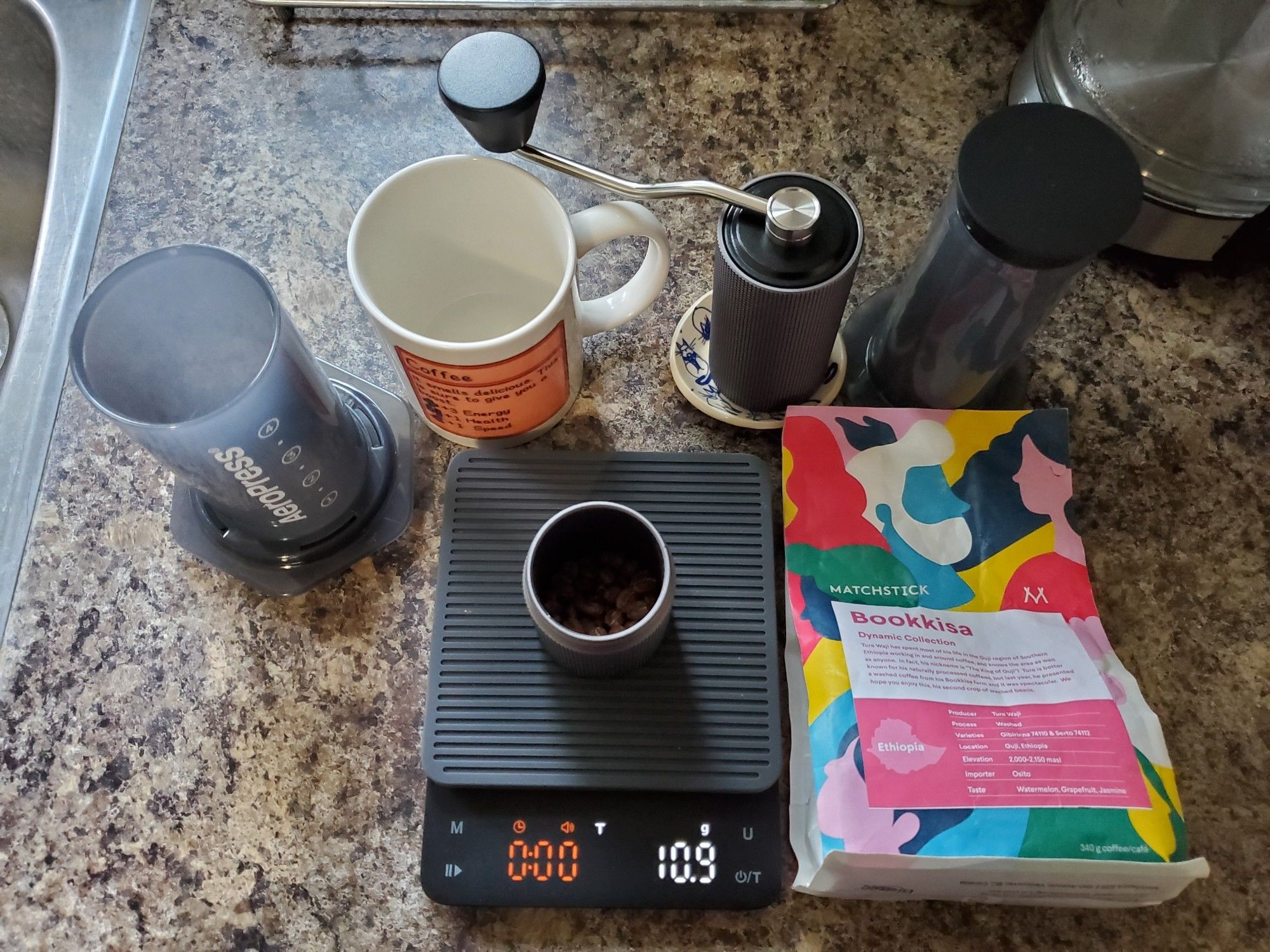 Coffee preparation setup: a scale, hand grinder, aeropress, and mug. A bag of coffee "Bookkisa" from Matchstick in also included.