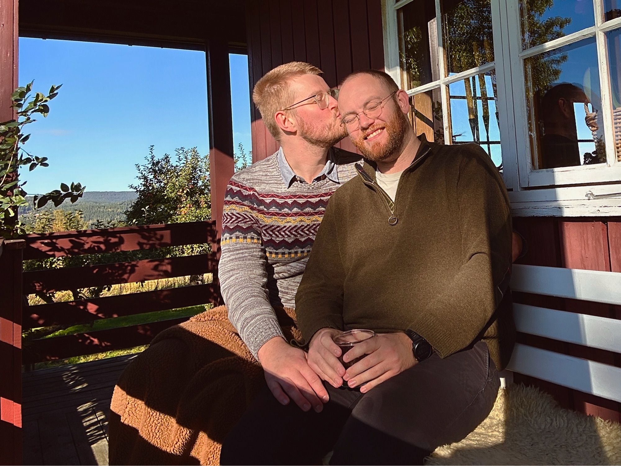 Man with glasses kissing man with glasses on head while sitting together on bench in sunlight