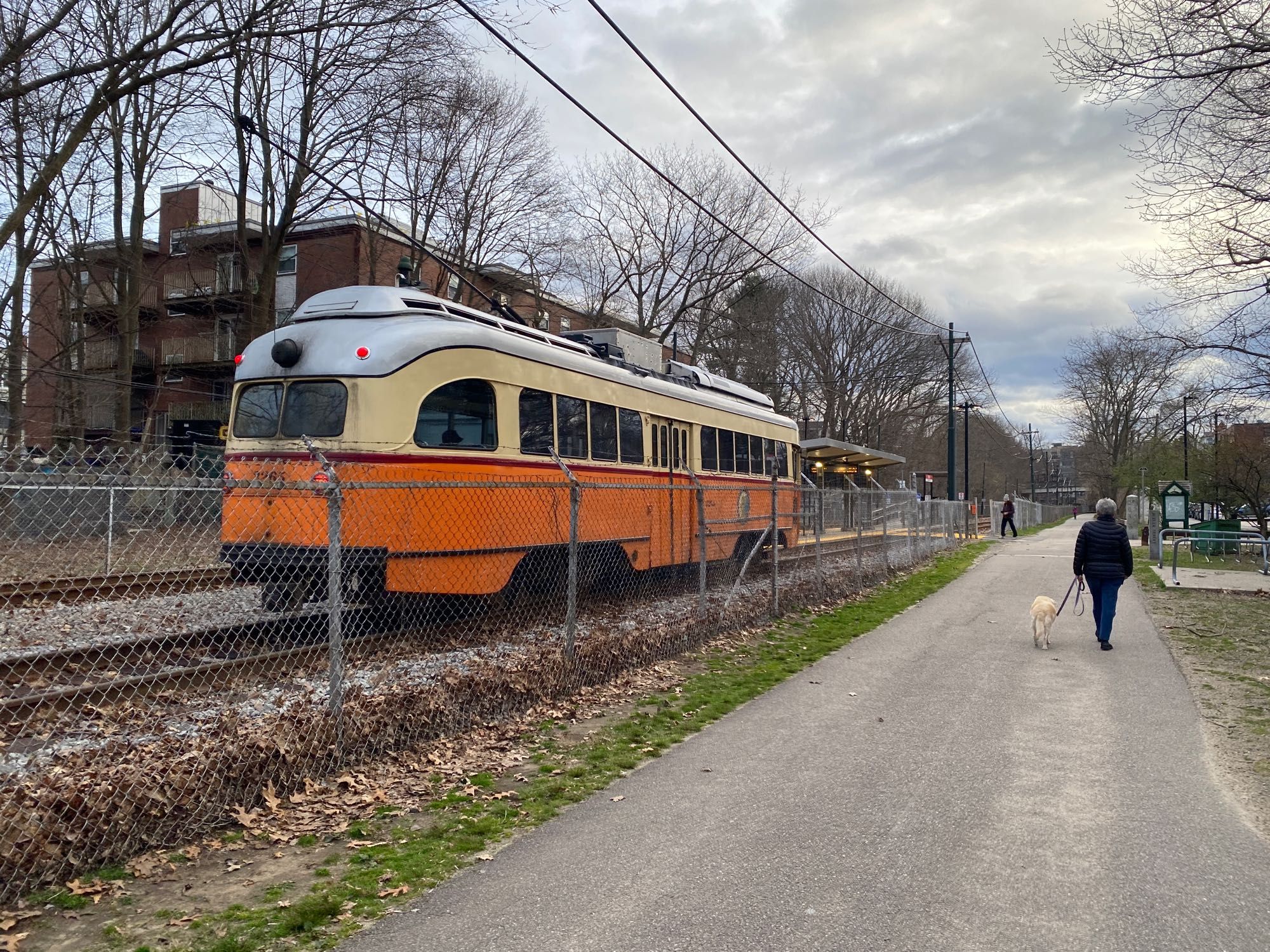 An orange MBTA trolley headed outbound approaching Butler Station in Dorchester, while a person walks their dog on the adjacent Neponset River Greenway RailTrail