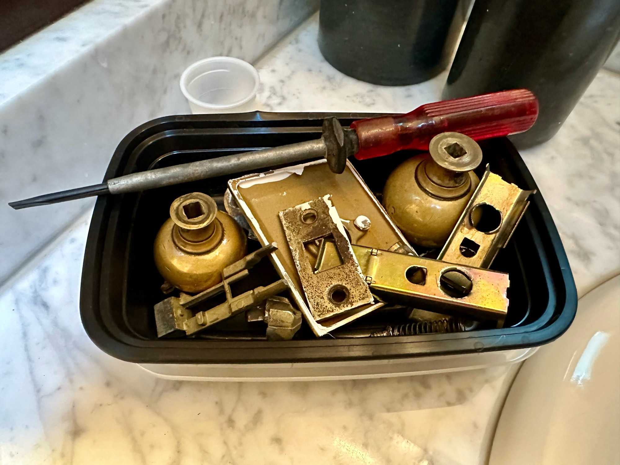 A black takeout container filled with the bits and pieces of an old brass doorknob and lock.