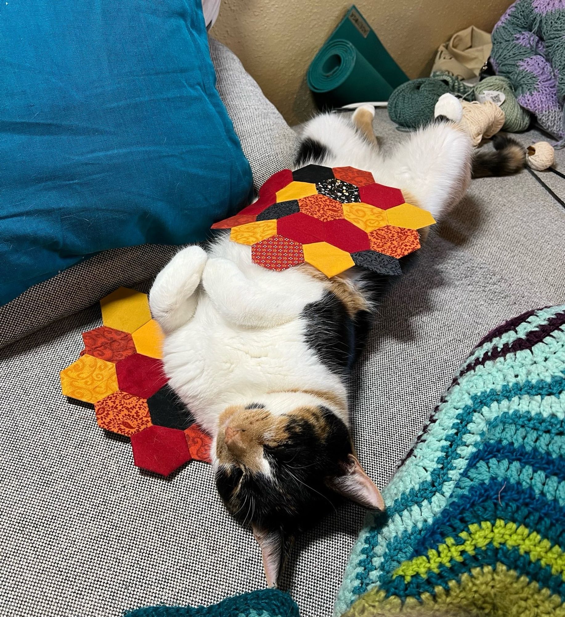 Calico cat lying on her back on a grey couch, resting atop a hexagonal quilt block in reds, yellows, and blacks, with another block lying on her belly.