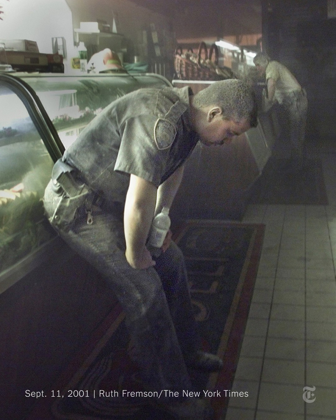 A police officer is bent over, coughing in a deli near the World Trade Center after the towers collapsed. His hands are on his knees, and he is covered in ash. Behind him, there is another man covered in soot. Photo by Ruth Fremson/The New York Times.