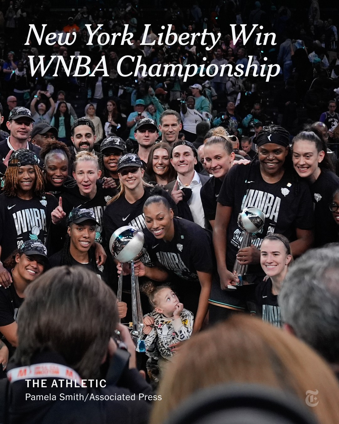 A group of baskeball players pose for a photo around a championship trophy. A child is in the front of the group. A headline reads "New York Liberty Win WNBA Championship." Photo by Pamela Smith/Associated Press 