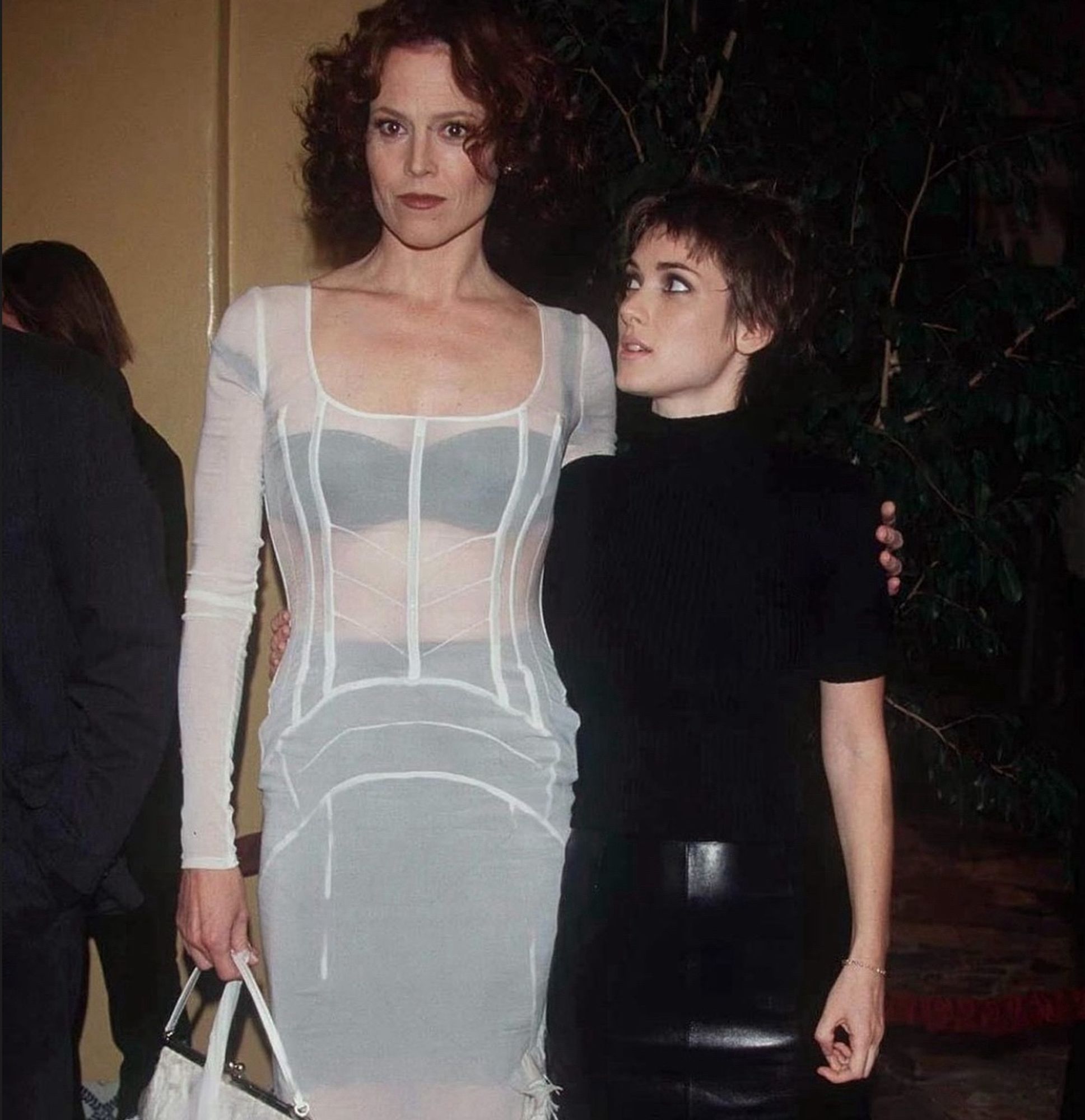 sigourney weaver and winona ryder - a tall white woman on the left with short dark hair in a white dress with her arm around a shorter white woman on the right with short dark hair, the woman on the right is gazing up at her while the woman on the left looks at cameras pointed at her