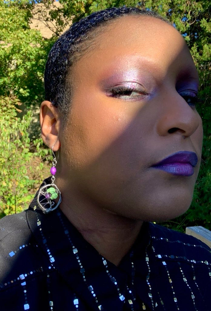 selfie of adrian modeling a dangly silver earring that features an amethyst bead connecting to titanium wire woven into a pentagram with two green howlite skull beads and an amethyst bead in the center of it.

he’s also wearing a purple smoky halo eye, purple lipstick and black button down top with silver sequins going down it like pinstripes. behind him are lush, green trees and he’s posed with a third of his face and adorned left ear in the sunlight while the other two thirds of his face are in the cool shade.