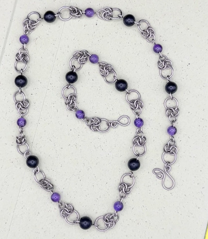 close up shot of the silver titanium chain necklace featuring a half byzantine weave with amethyst and onyx beaded links. the weave features an alternating pattern of 3 interlocking small loops, connected to slightly bigger rings in between round onyx and amethyst beads.