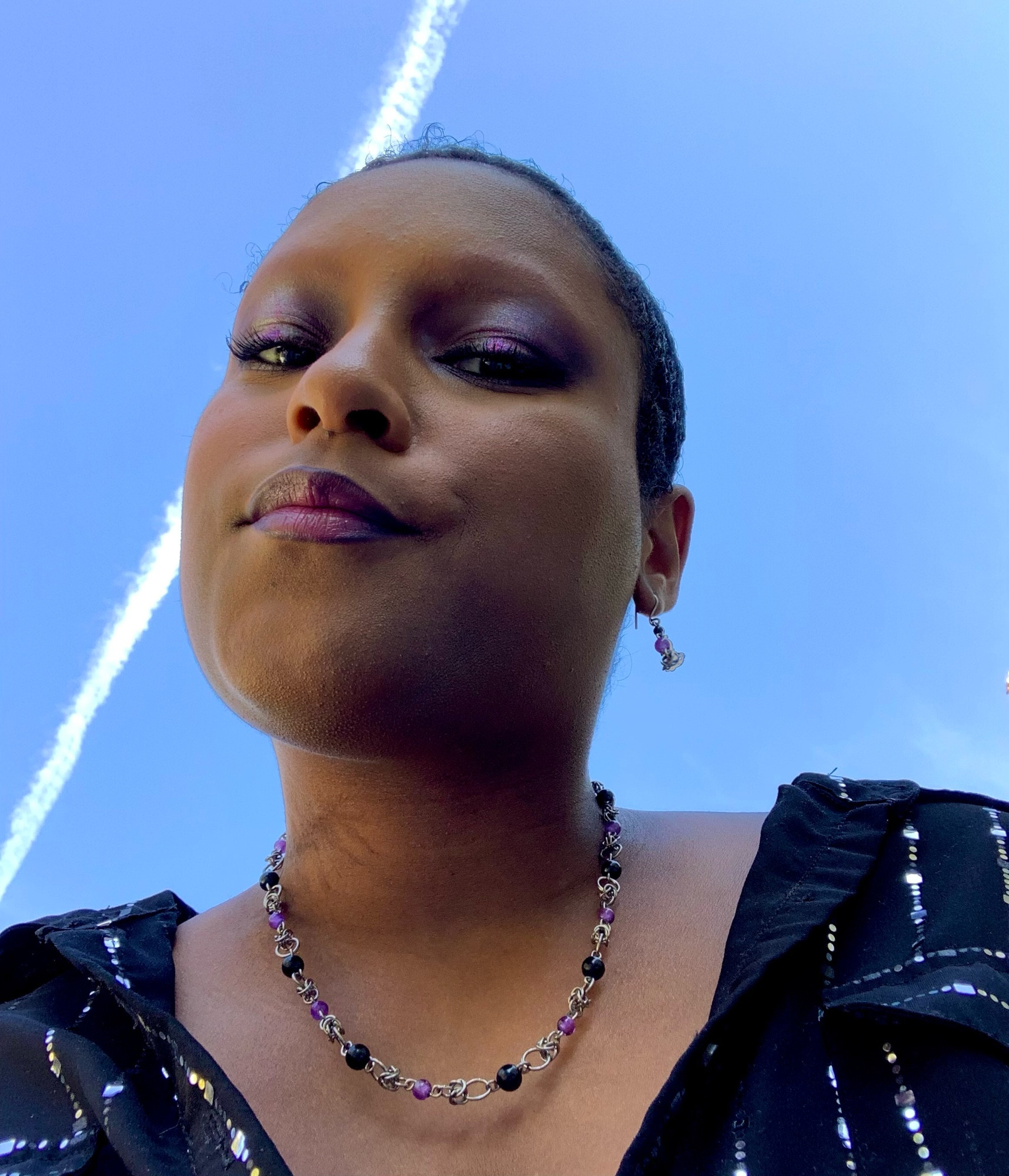 selfie of adrian modeling 2 pieces of jewelry: a pair of titanium earrings featuring beaded onyx and amethyst links and titanium chain necklace featuring a half byzantine weave with amethyst and onyx beaded links.

he’s also wearing a purple smoky halo eye, purple lipstick and black button down top with silver sequins going down it like pinstripes. 

adrian is posed looking down at the camera with the bright blue sky behind him and a plane streak going across his left side.