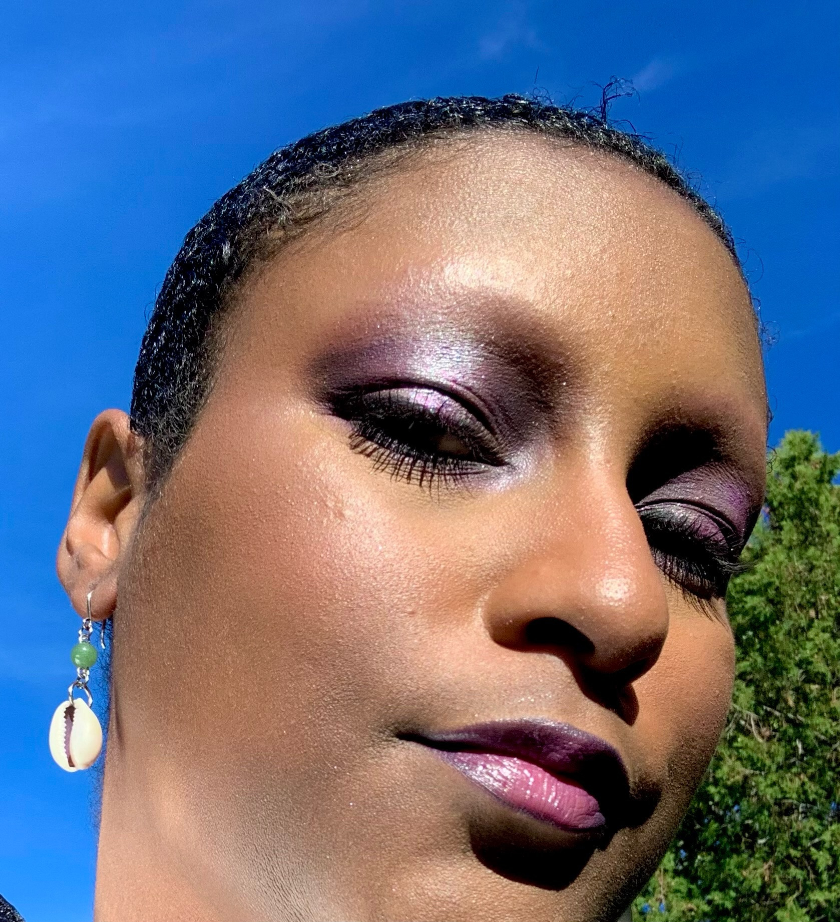 selfie of adrian modeling a dainty silver titanium earring that features a green aventurine bead and cowrie shell at the end.

he’s also wearing a purple smoky halo eye, purple lipstick and has a bright blue sky and tree leaves behind him.