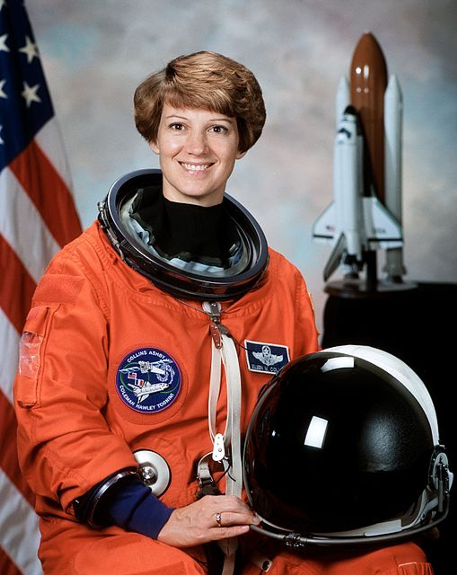 Eileen Collins in her official mission photo. She is a white woman with red hair, wearing her orange spacefliht suit and holding the helmet. A model of the space shuttle is on a desk behind her.