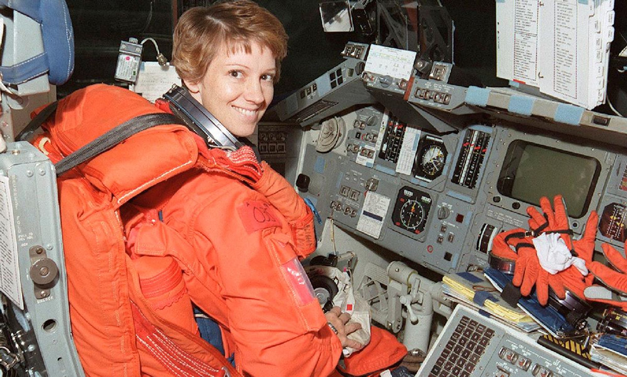 Eileen Collins in her orange launch suit, seated at the controls of Columbia in 1999. She's a white woman with red hair.