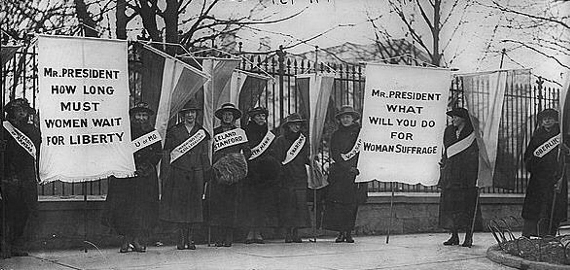 Nine of the silent sentinels. All are white women wearing sashes and some are holding up banners reading 'Mr President: how long must women wait for liberty", 'Mr President: what will you do for Woman suffrage"