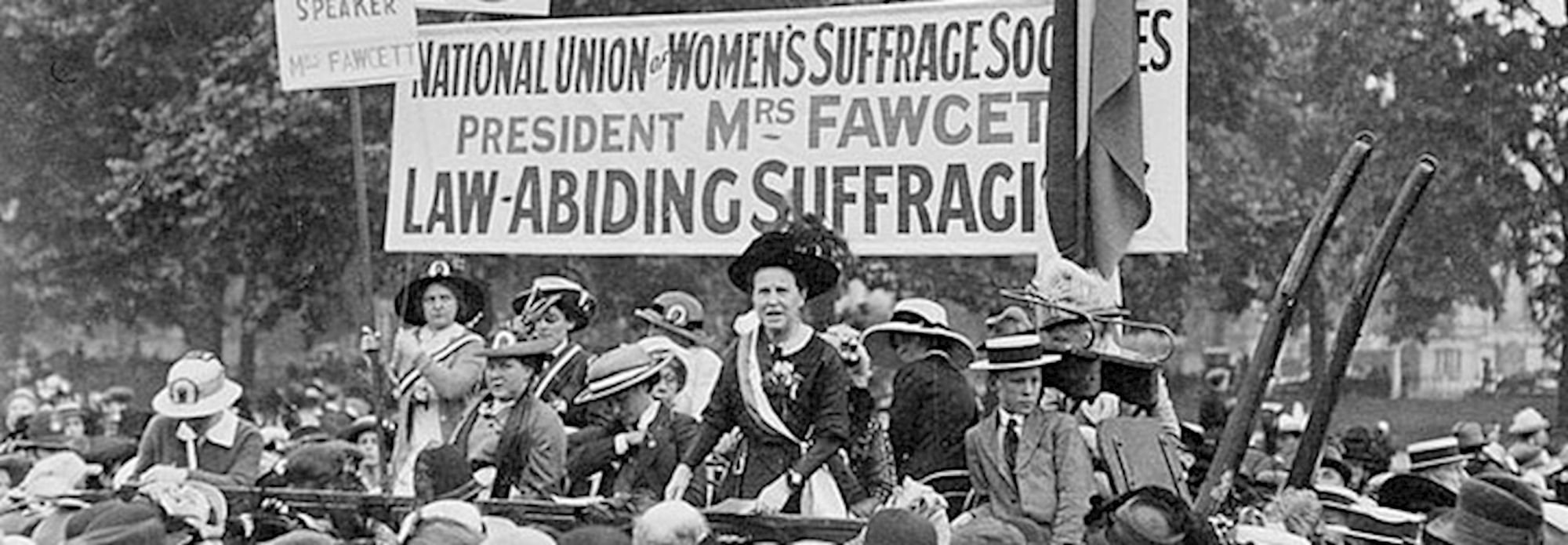 Millicent Fawcett addressing the crowd at Hyde Park. The banner behind her highlights that her National Union of Women's Suffrage Societies are "law abiding suffragists". All the women on the speaking platform are white.