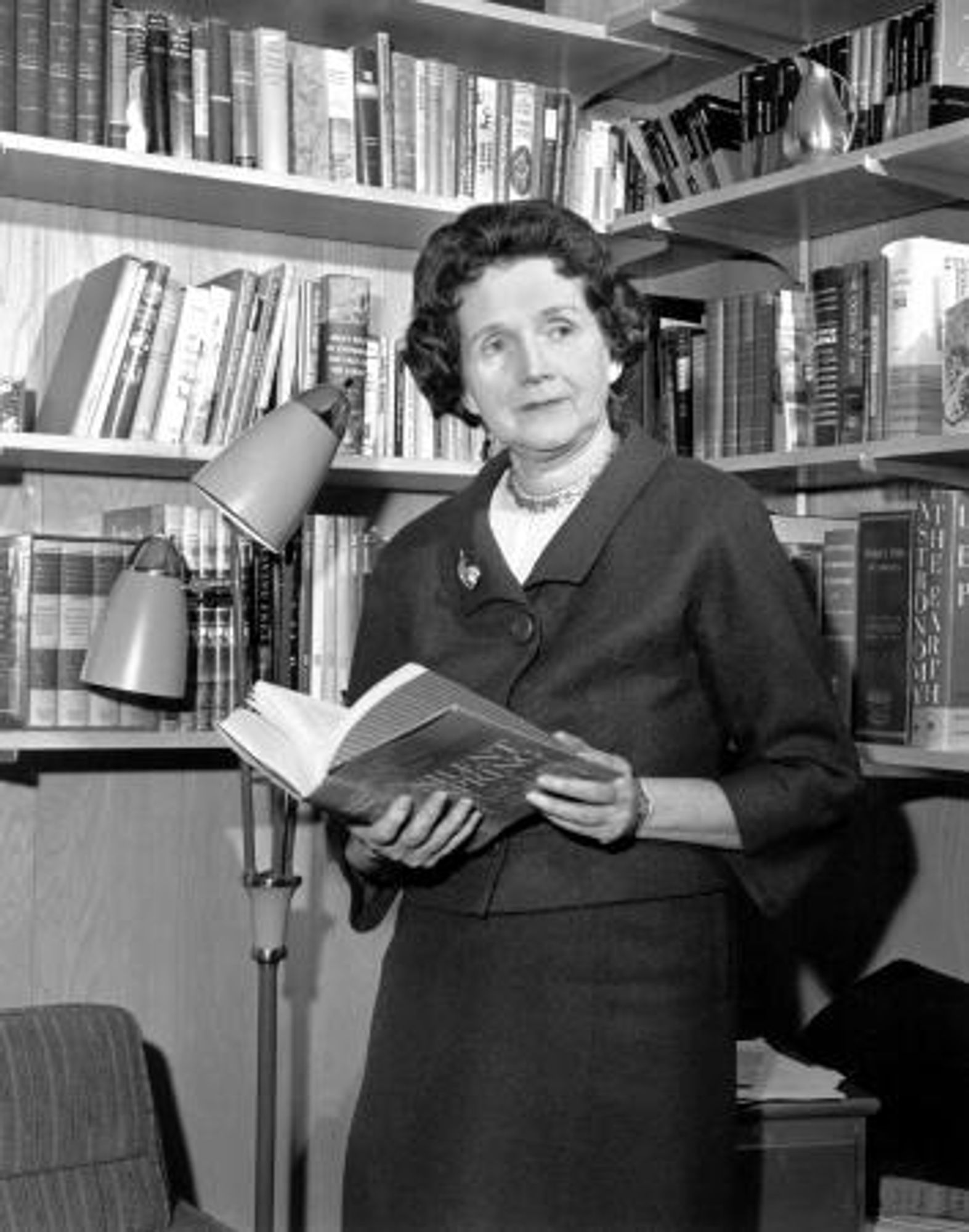 Rachel Carson with a copy of Silent Spring. She is a white woman with dark hair standing in front of a bookcase.