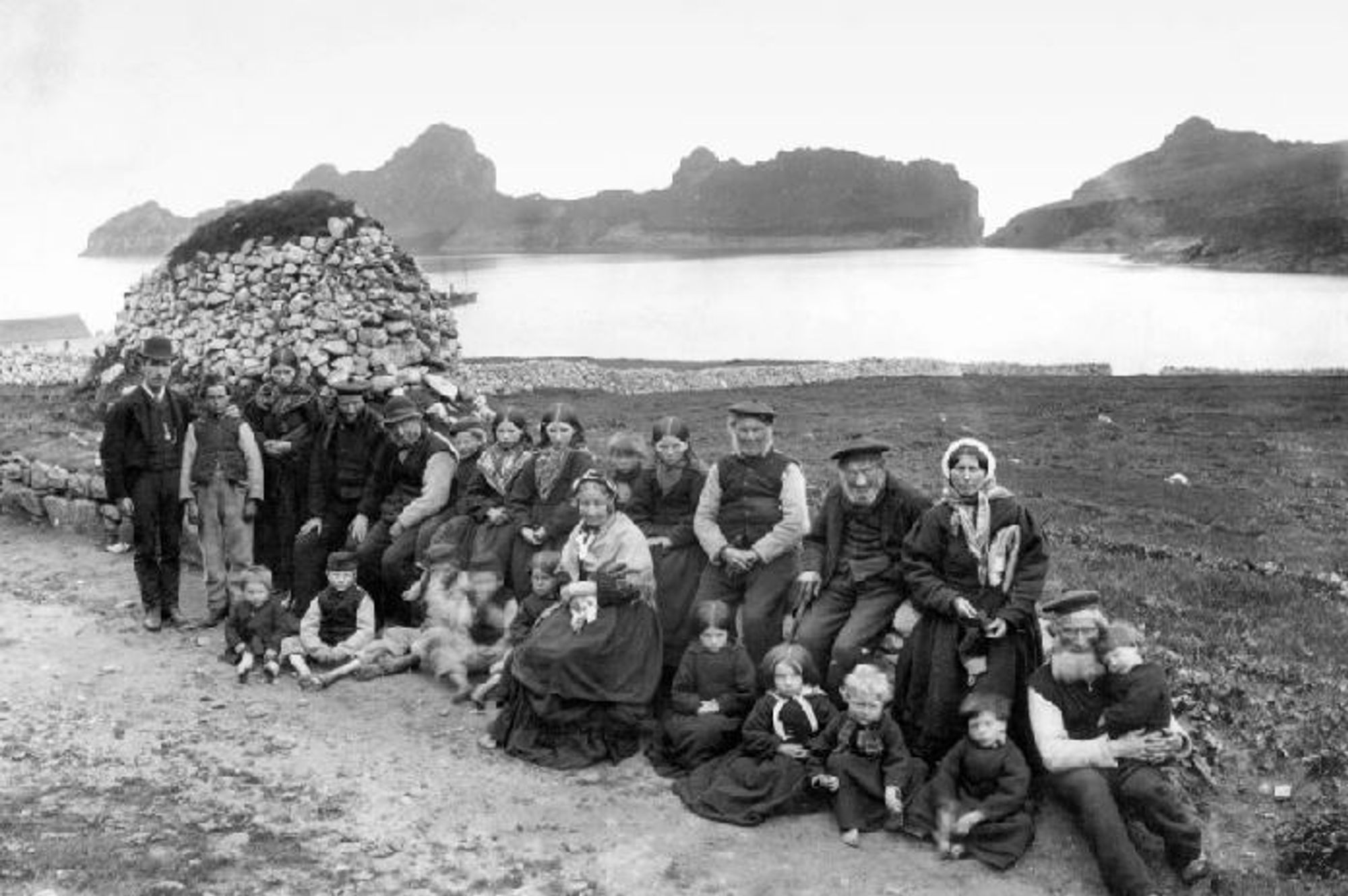 A group of the St Kildans. The women and old men are sitting on a wall and the children on the ground.
