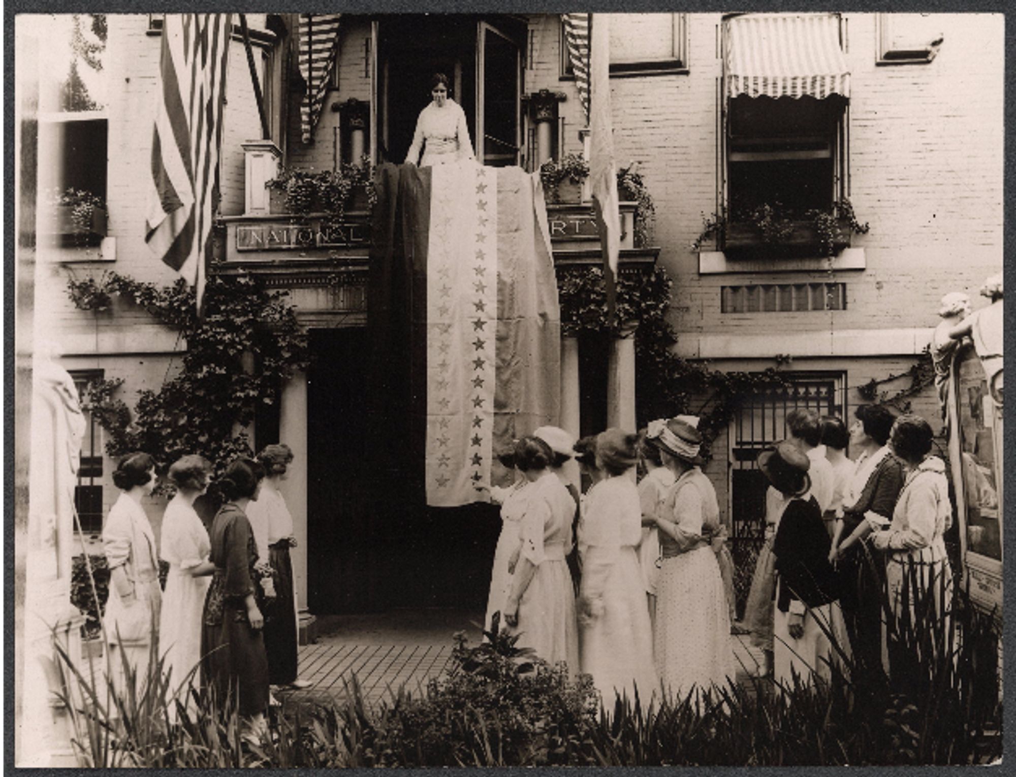 Alice Paul standing on a balcony with a flag showing a star for each state that has ratified the amendment. A group of women stand below it. Everyone is white.