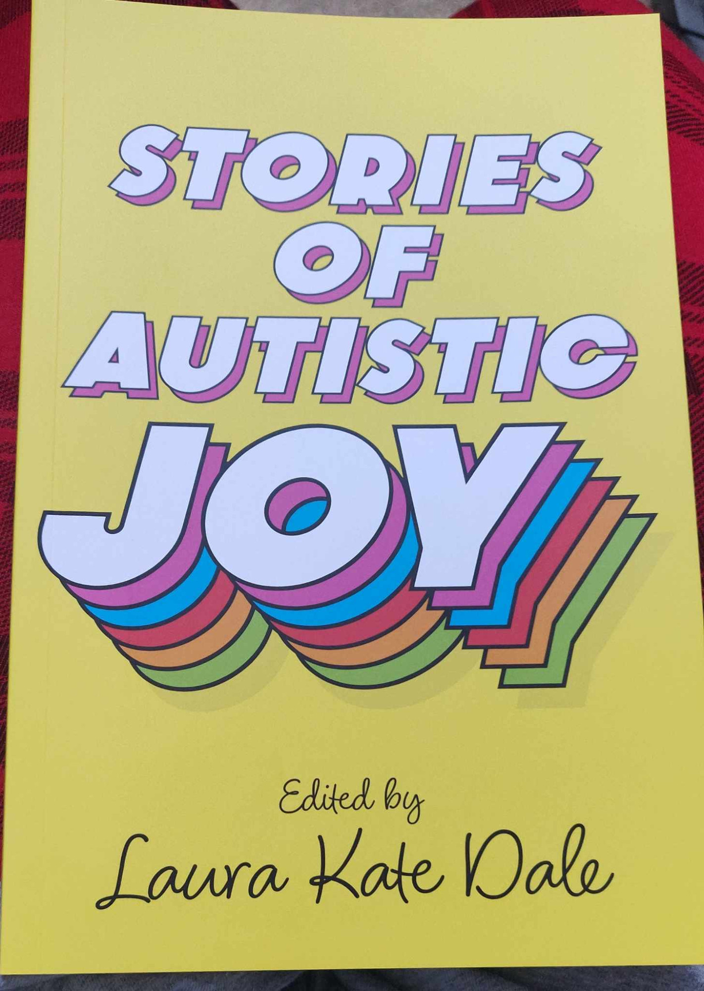 The front cover of the book reads Stories of Autistic Joy, edited by Laura Kate Dale. The word joy is layered multiple times in different bright colours.