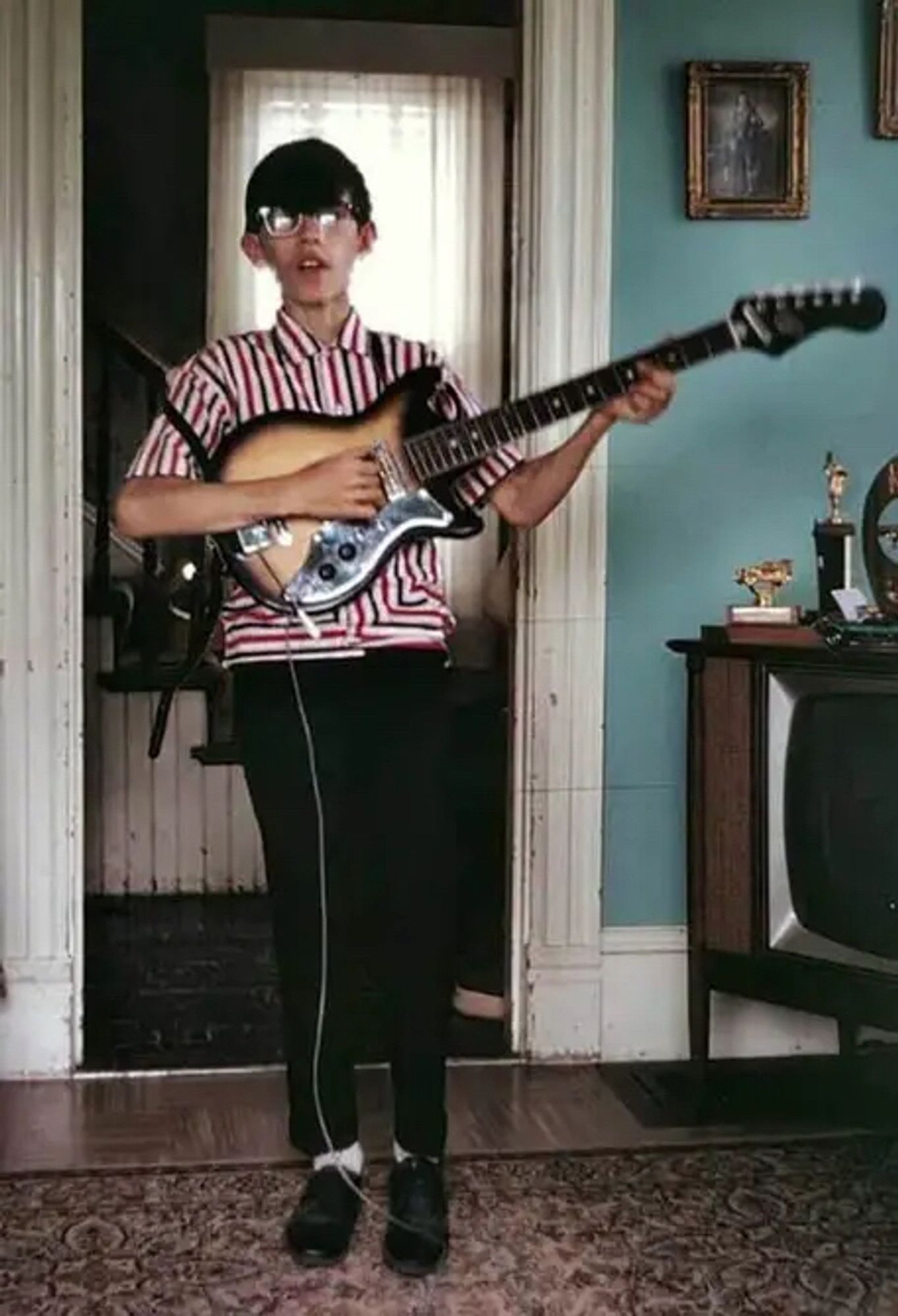 A skinny young lad with short black hair, glasses. Wearing a striped shirt, holding an electric guitar. An old TV set can be seen off to the right. Teal coloured walls.