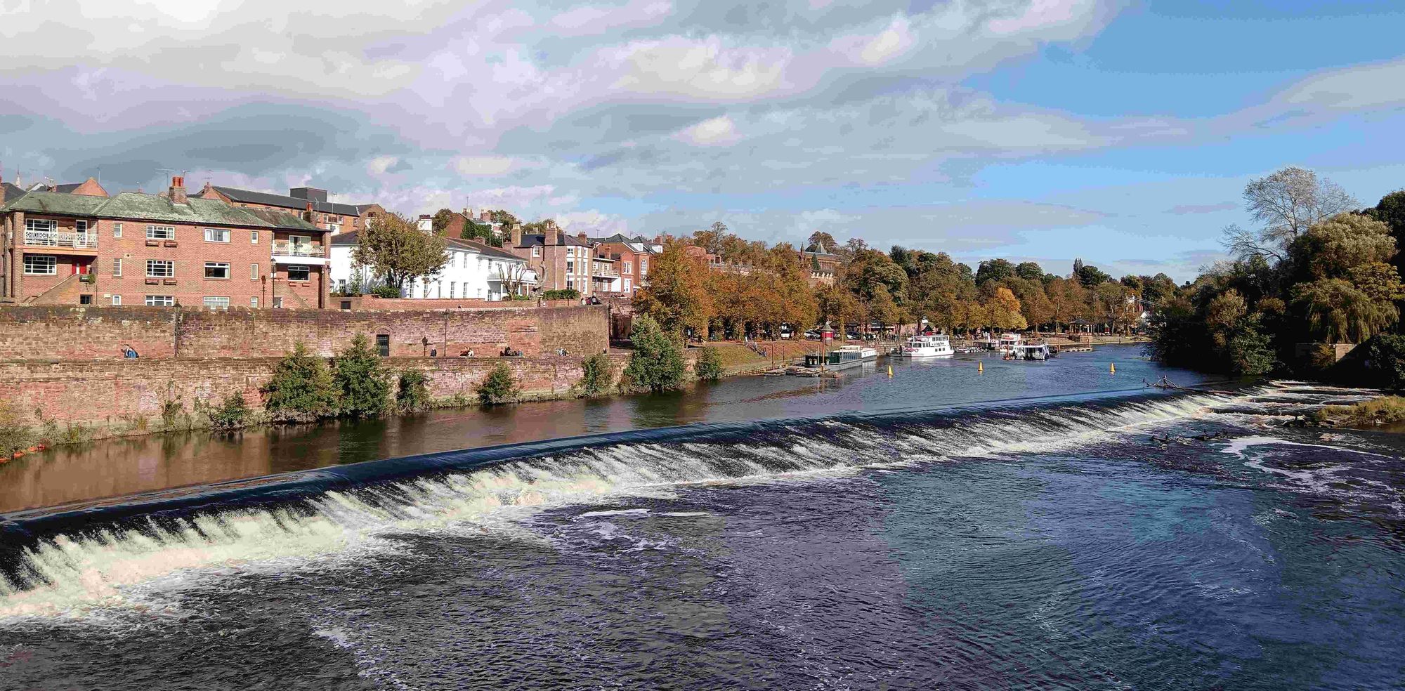 A weir runs diagonally across a broad river. Buildings line the left hand bank.