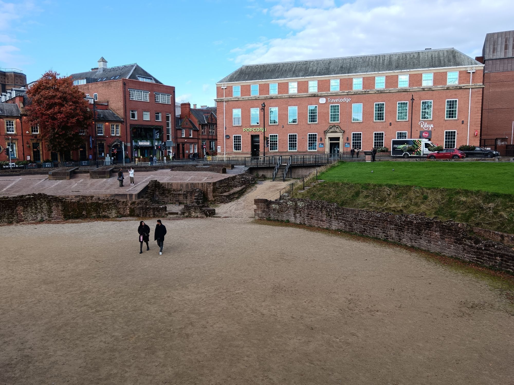 Part of an excavated Roman amphitheatre showing the main entrance. Modern buildings in the background.