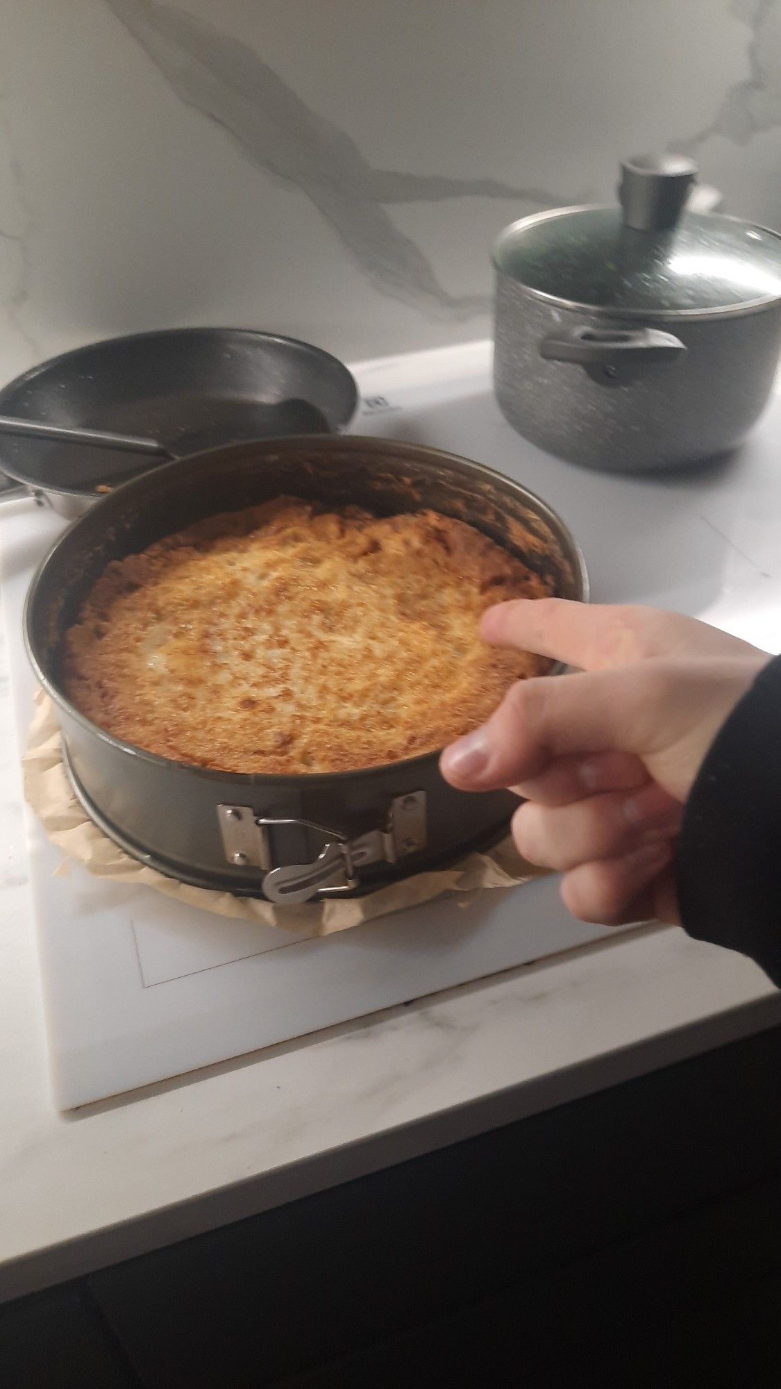 the image shows a person pointing at a metalic pan tray surrounding a pie atop a white induction stove, a few pots and pans on the stove as well