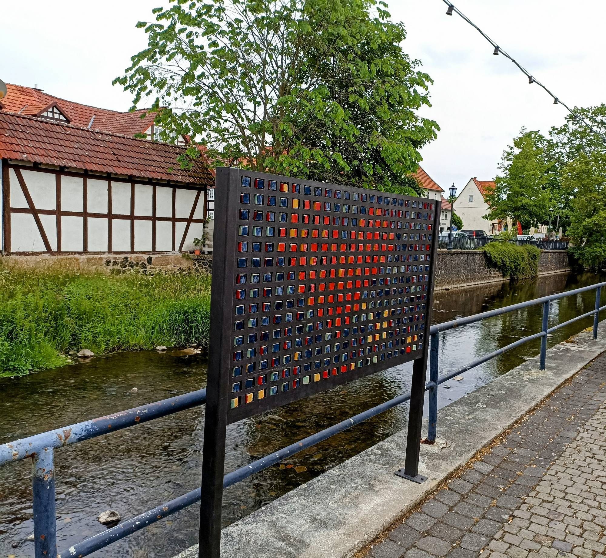 Bachlauf in einer Ortschaft, im Vordergrund eine Tafel mit einem Herz aus Mosaiksteinen