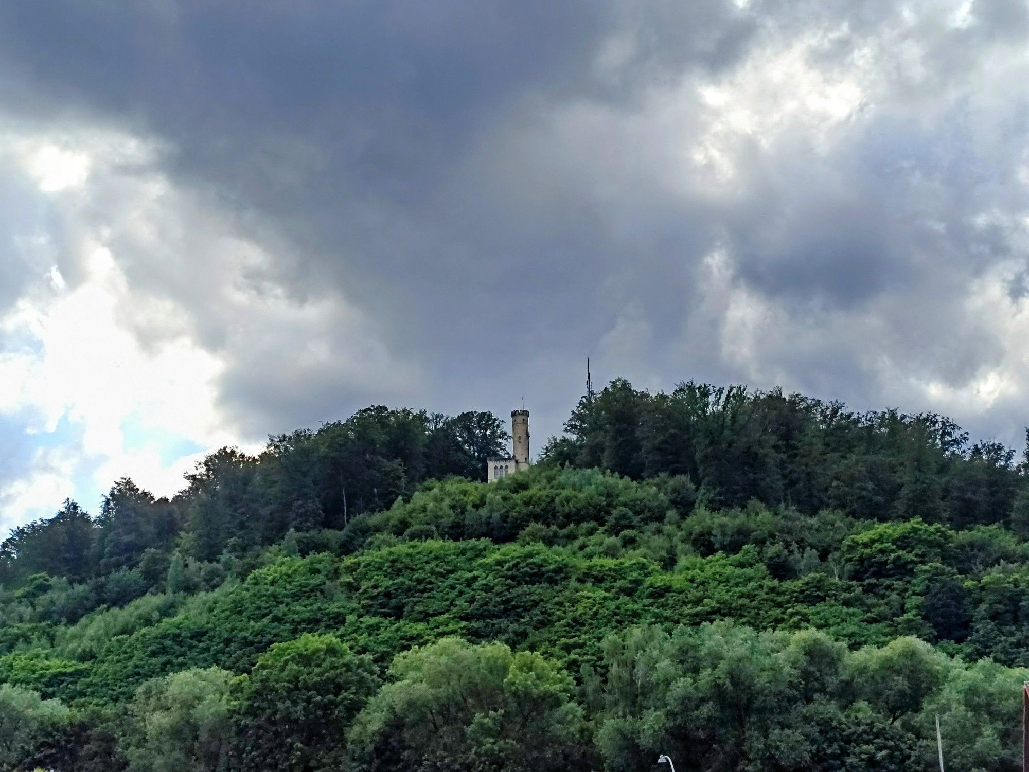 ein bewaldeter Berg, oben ein Turm, dramatische Bewölkung