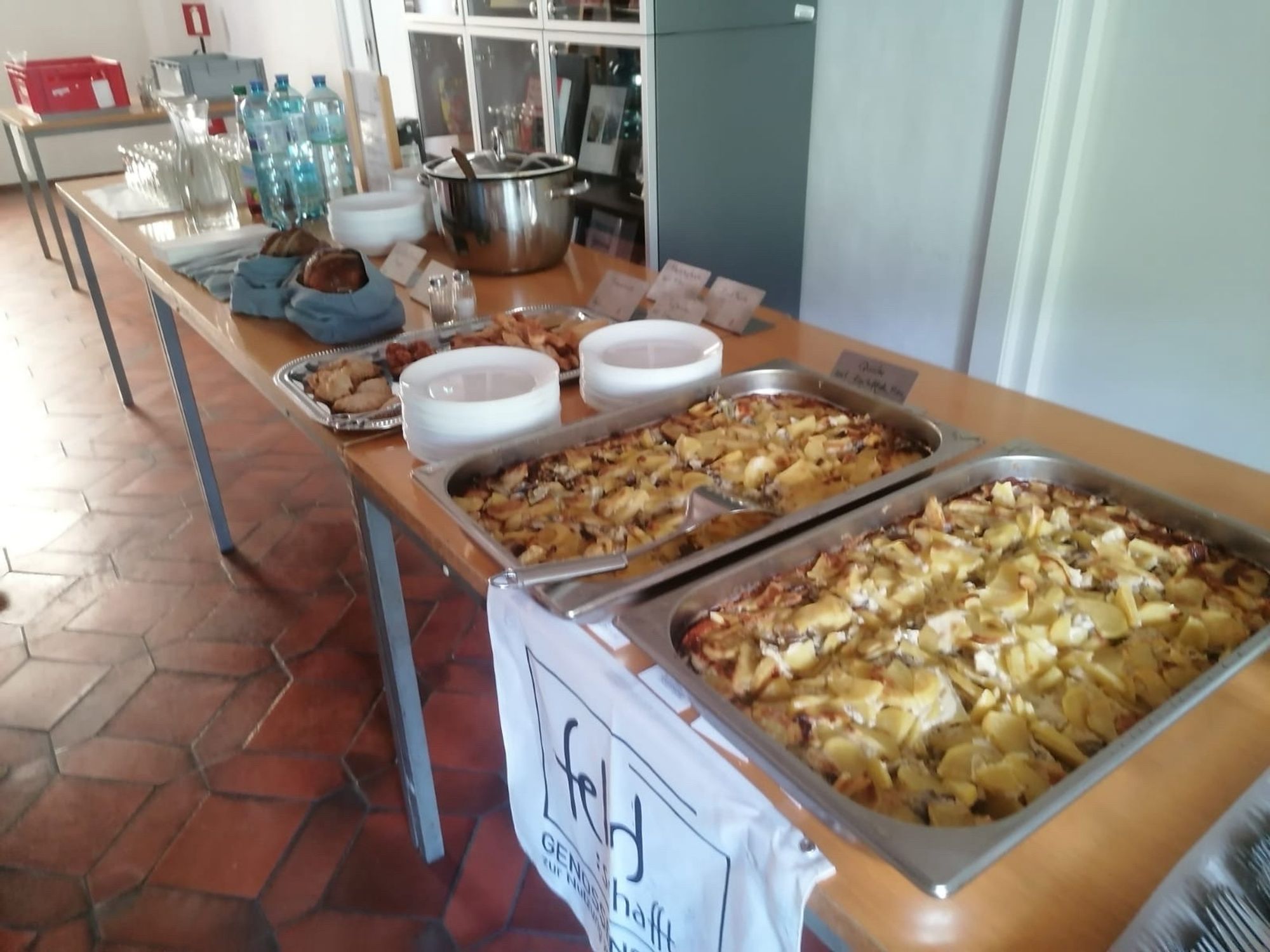 Ein langer Tisch mit zwei Metallbehältern mit einem Pilz-Kartoffel-Auflauf. Im Hintergrund Brot, Gläser, Wasser, Saft und Kaffee.